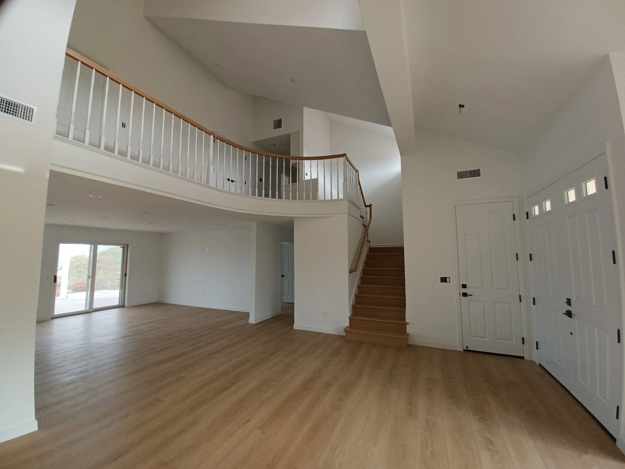 Empty living room in a newly built house with wooden floors, white walls, a staircase with wooden steps and railing, sliding glass doors, and a high vaulted ceiling.