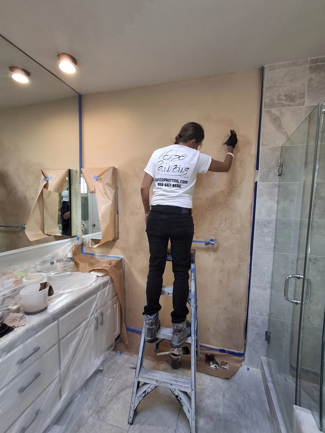 A person standing on a ladder painting a beige wall in a bathroom, with a shower on the right and a white vanity with a sink on the left, protected with paper and painter's tape. Faux paint in Mission Beach California.