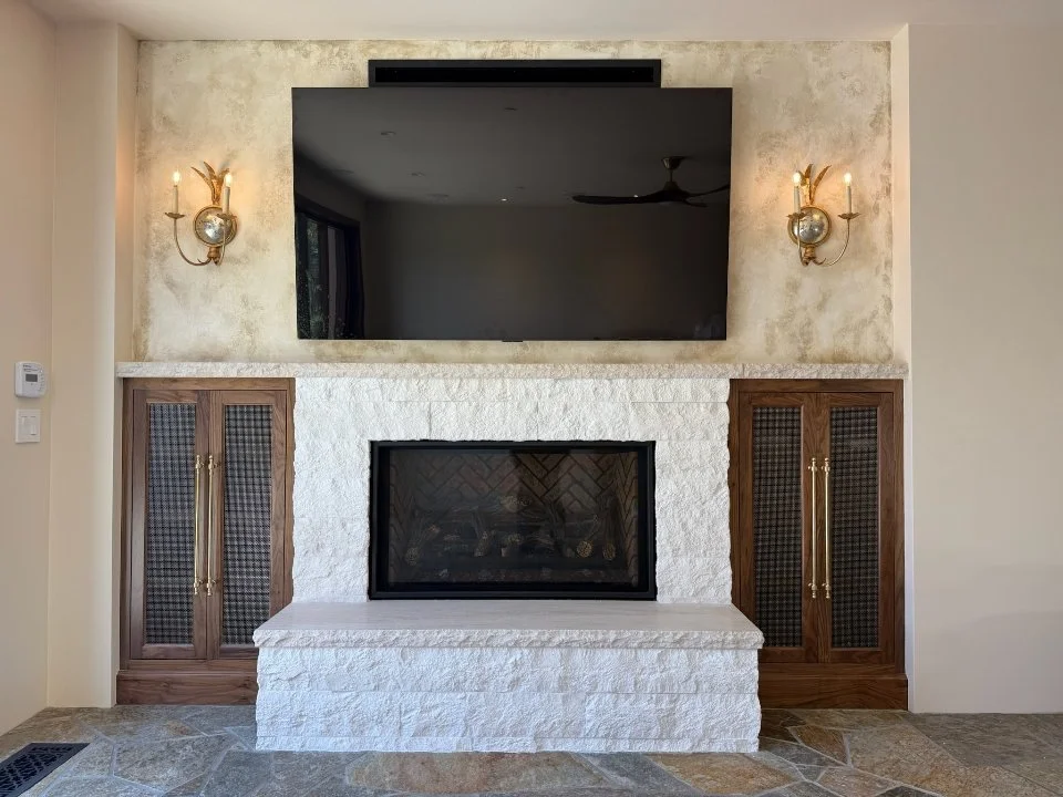 Living room with a white stone fireplace, a mounted flat-screen TV above it, and two wooden cabinets with gold handles on either side, illuminated by wall sconces.