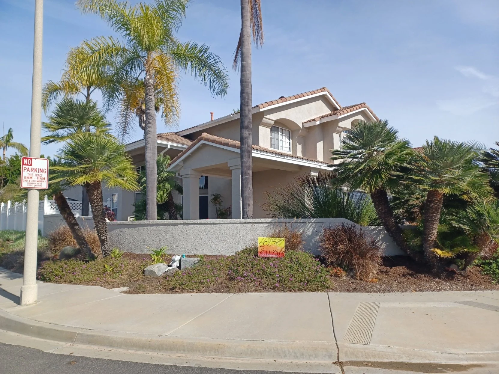 A two-story suburban house with a beige exterior and terracotta roof tiles, surrounded by palm trees and desert plants, with a sidewalk and street in the foreground.