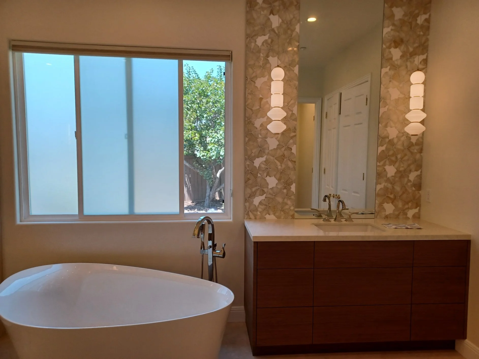 Bathroom with a large frosted window, a freestanding bathtub, a vanity with a sink, a large mirror, and decorative wall tiles with a flower pattern. There are two pendant lights on either side of the mirror and a view of greenery outside.