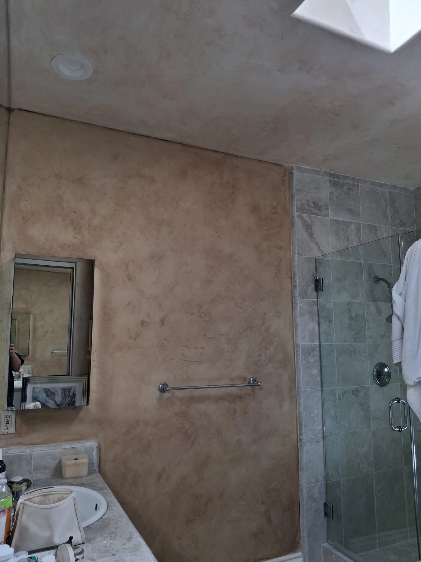 A bathroom with a beige textured wall, a shower with a glass door and tiled walls, a mirror above the sink, and various toiletries on the counter. Fauz paint in mission beach california.