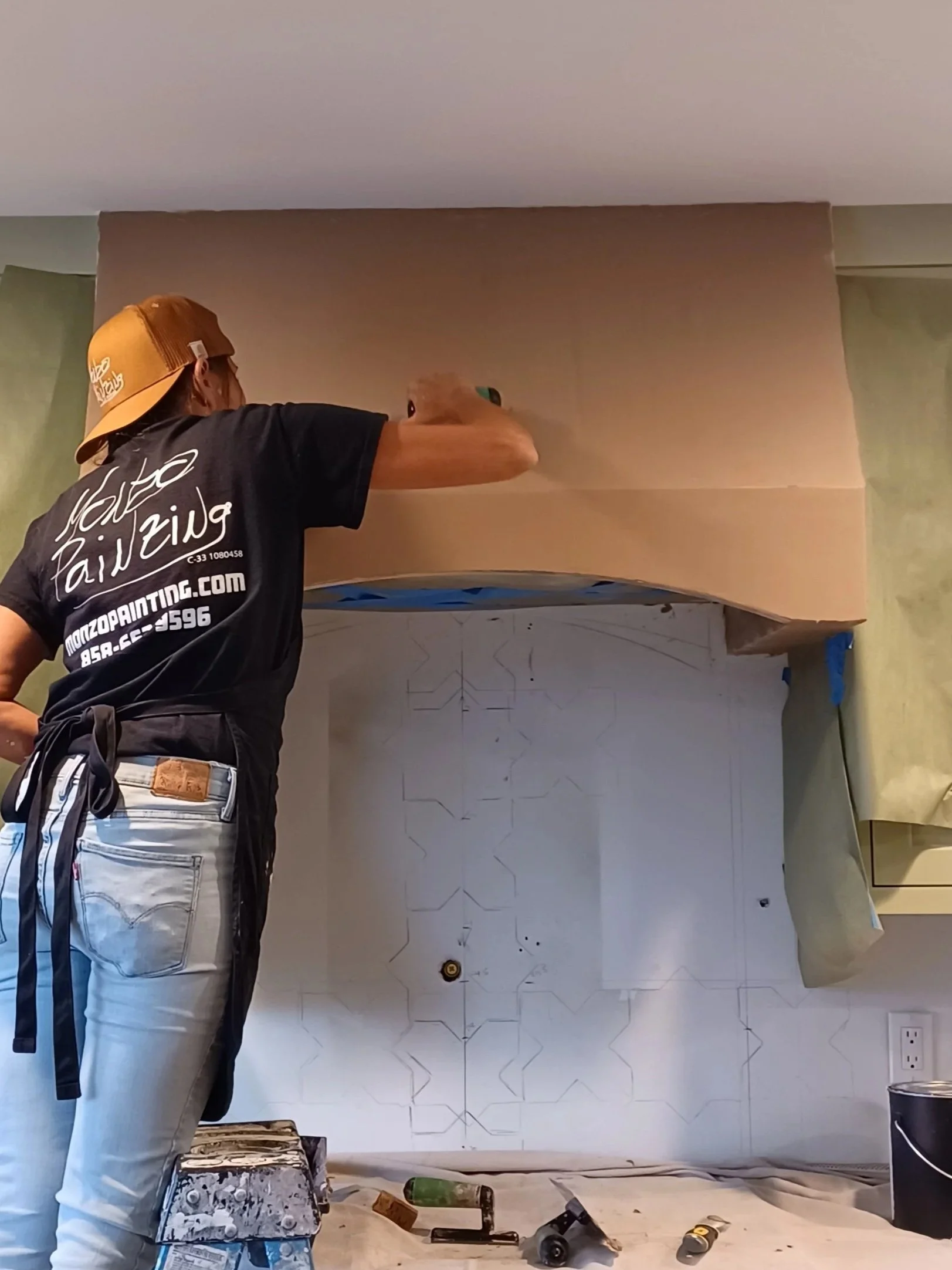 Person painting a wall with geometric patterns, wearing a black t-shirt and a beige cap, tools scattered around.