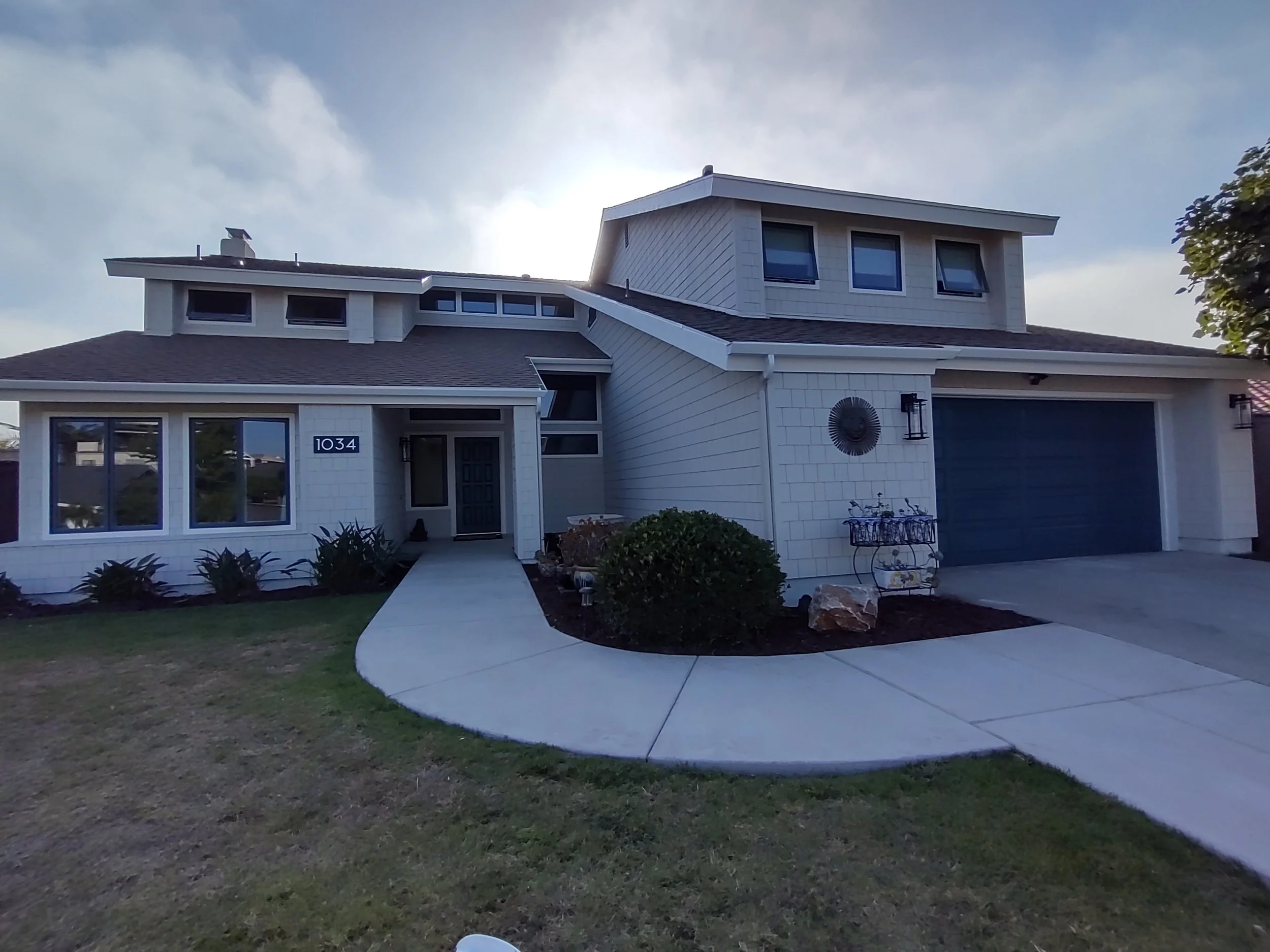 Modern two-story house with white exterior, gray roof, large front windows, decorative garden with plants and rocks, driveway, and a blue garage door.