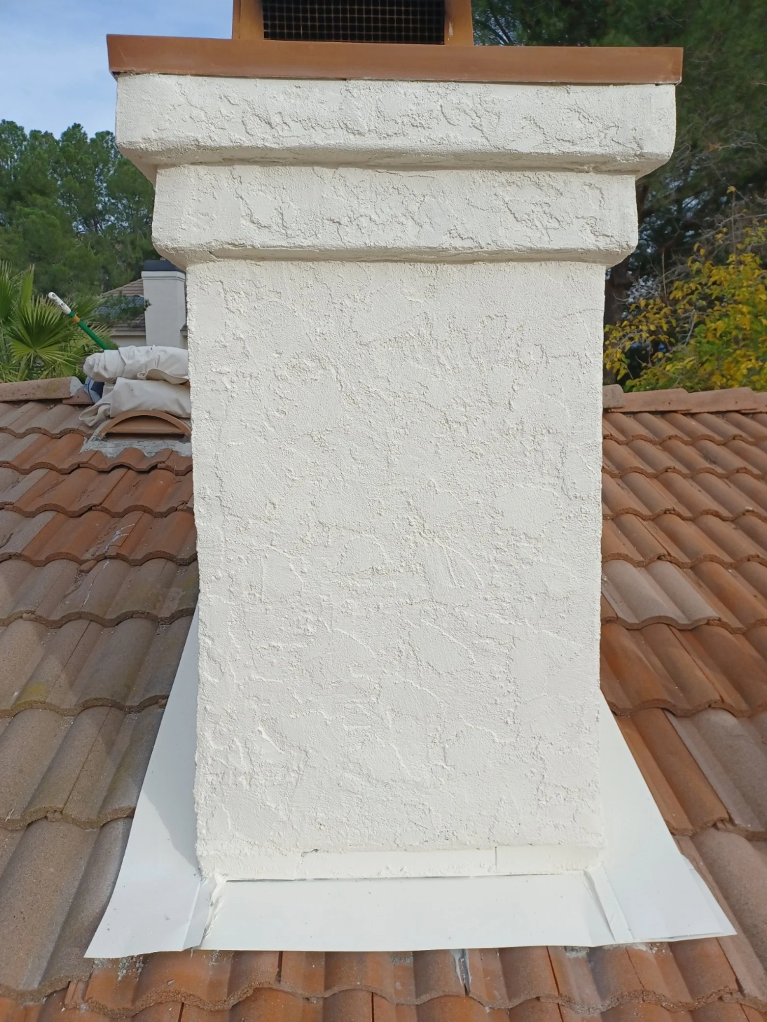 Close-up of a chimney with a textured white stucco finish, on a roof with terracotta tiles, and a background of trees and a partly cloudy sky.