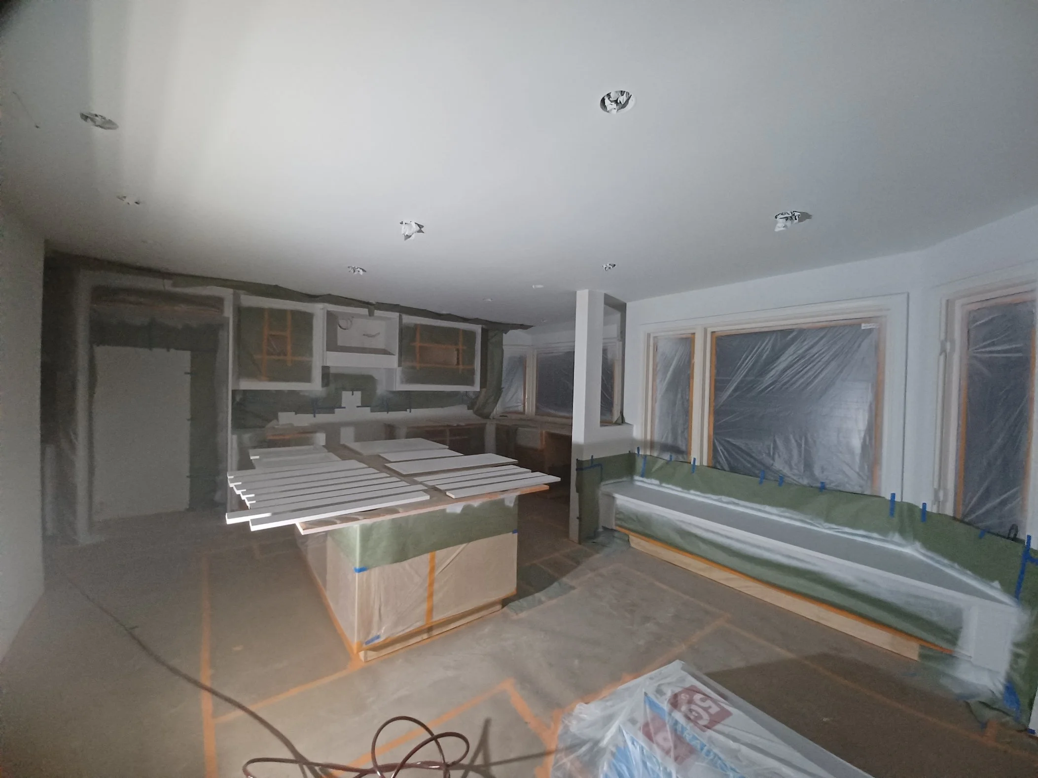 Room under renovation with plastic-covered furniture, unpainted walls, and holes in the ceiling, indicating ongoing construction or repair work.