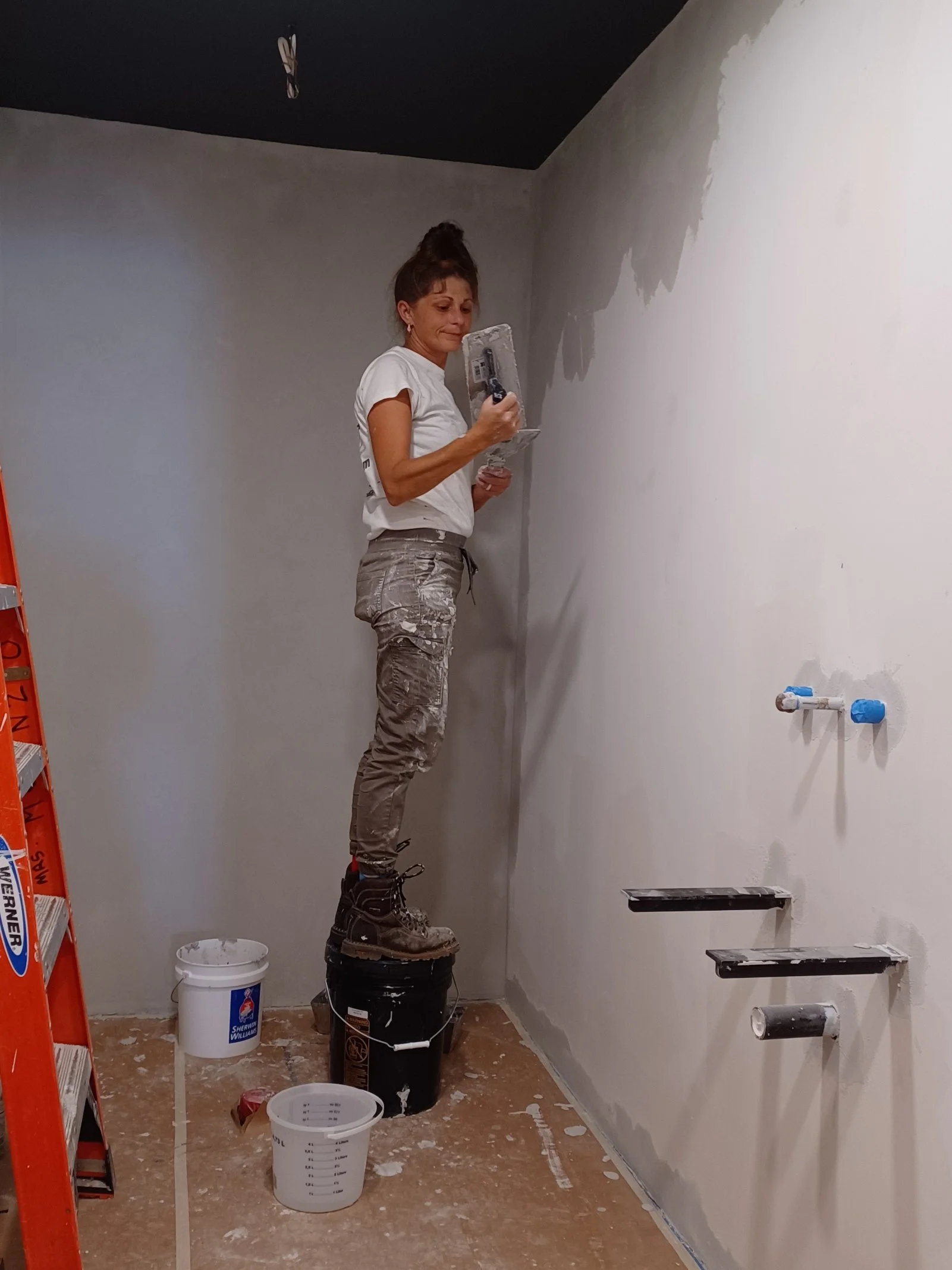 A woman standing on a bucket and painting a wall with a roller in a room under construction, with buckets, paint, and ladders around her.