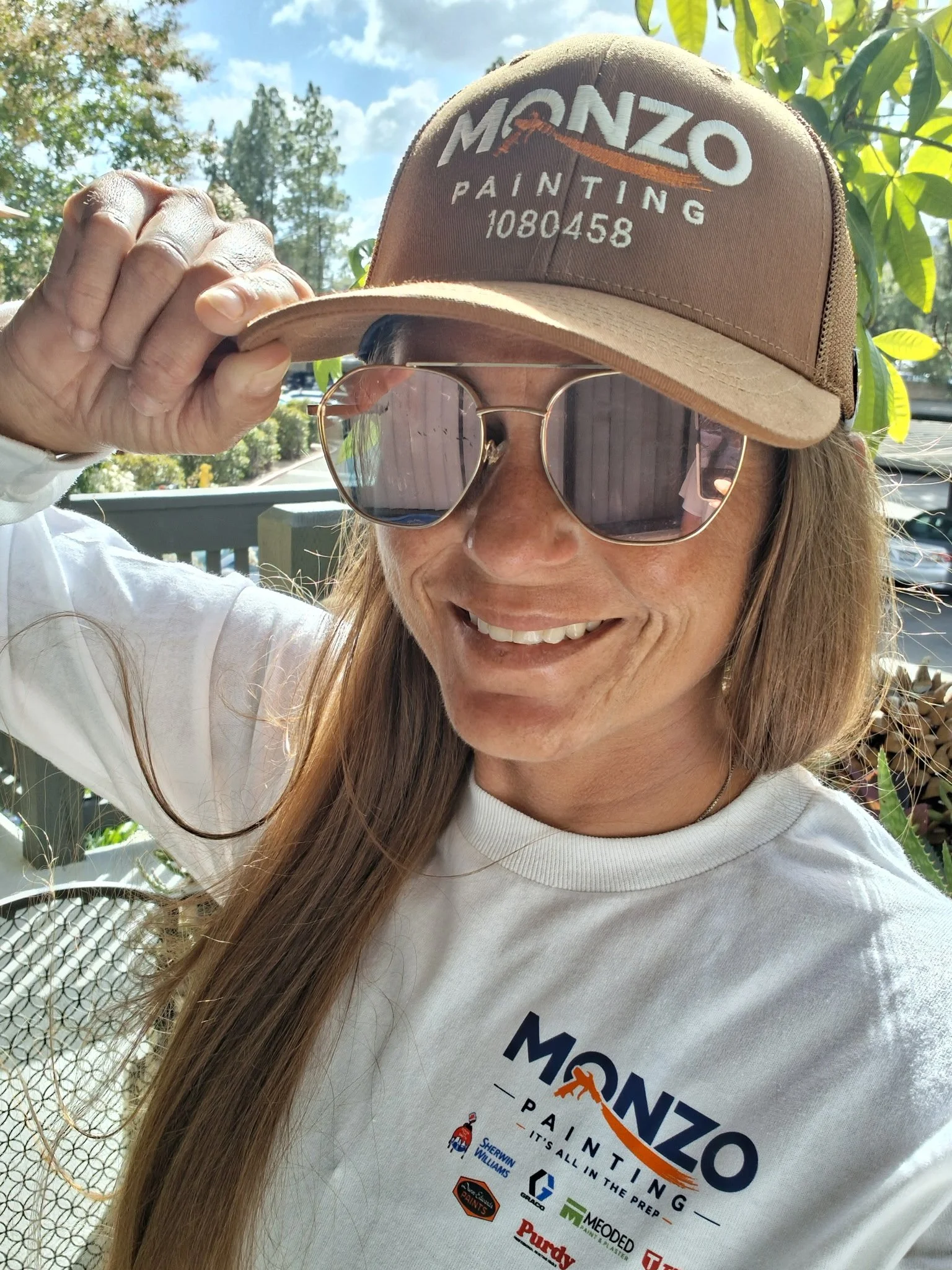 A woman wearing sunglasses and a beige cap with "Monzzo Painting" logo, adjusting the cap with her right hand, wearing a white monzo painting t-shirt, smiling outdoors with trees and a blue sky in the background.