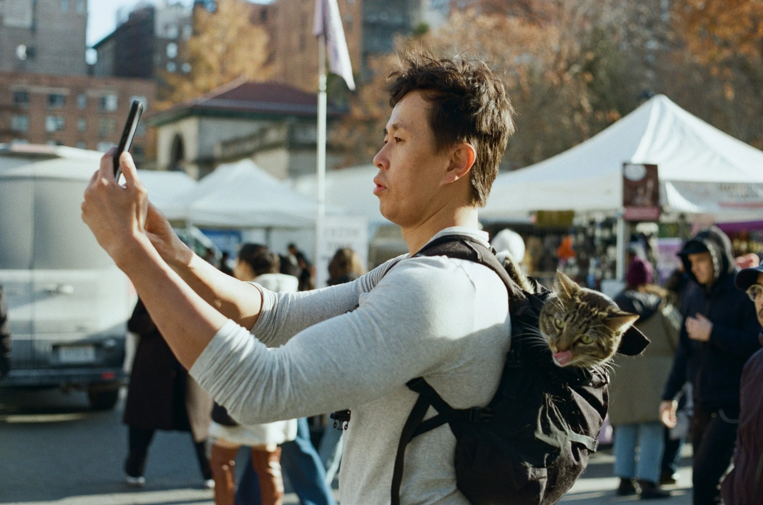 Playful analog film street photograph of a man photographing on his phone with a cat peeking out of his backpack at an outdoor market in New York City, shot by Dario Tapia.