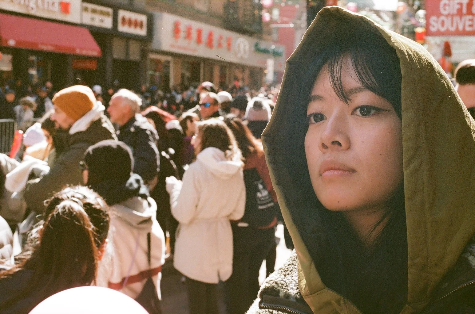Intimate analog film portrait of a young woman in a hooded jacket amid a crowded Chinatown street in New York City, captured by film photographer Dario Tapia.