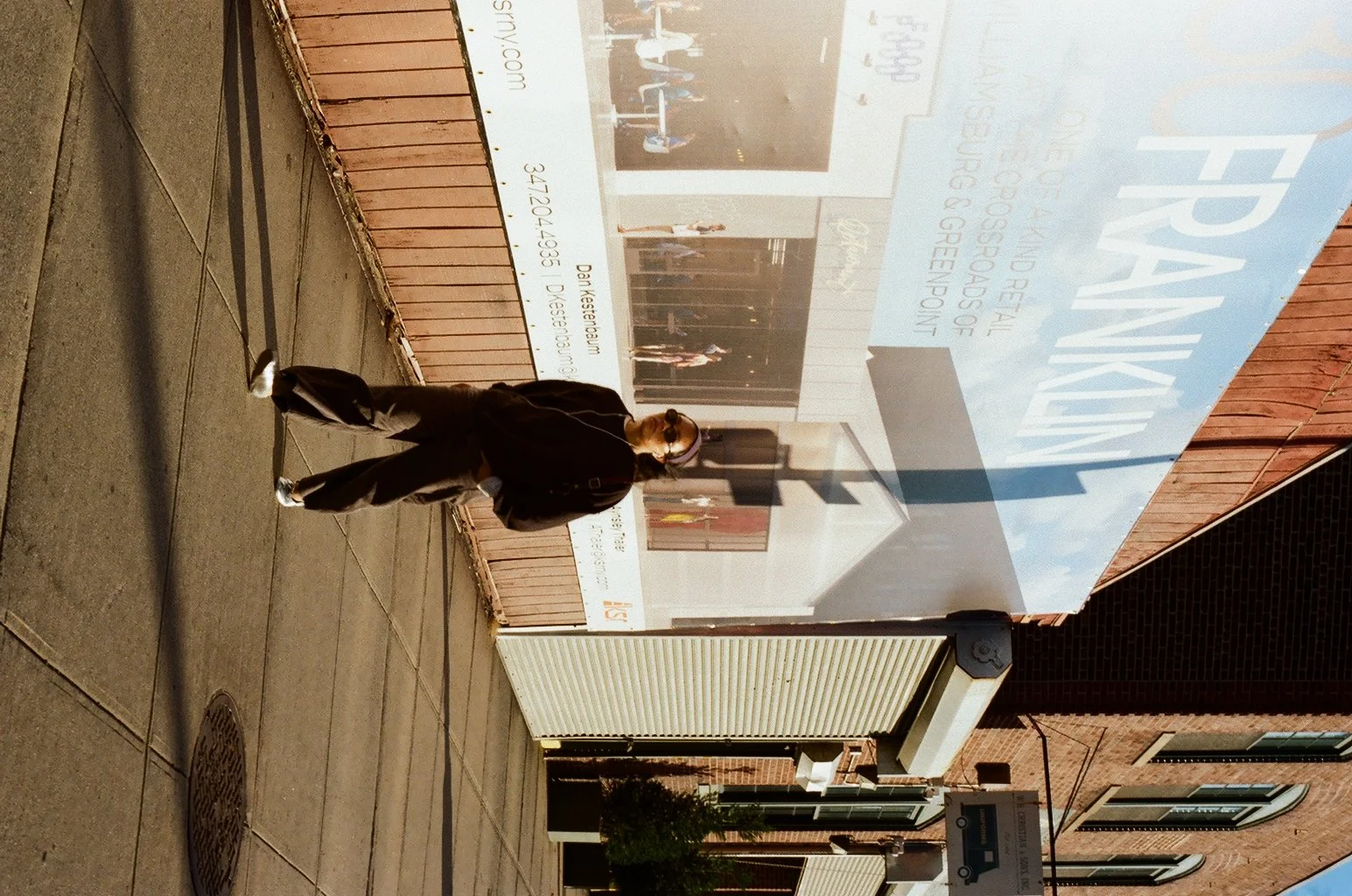 Analog film street photograph of a lone figure on a Brooklyn sidewalk beneath a large billboard, captured by NYC film photographer Dario Tapia.