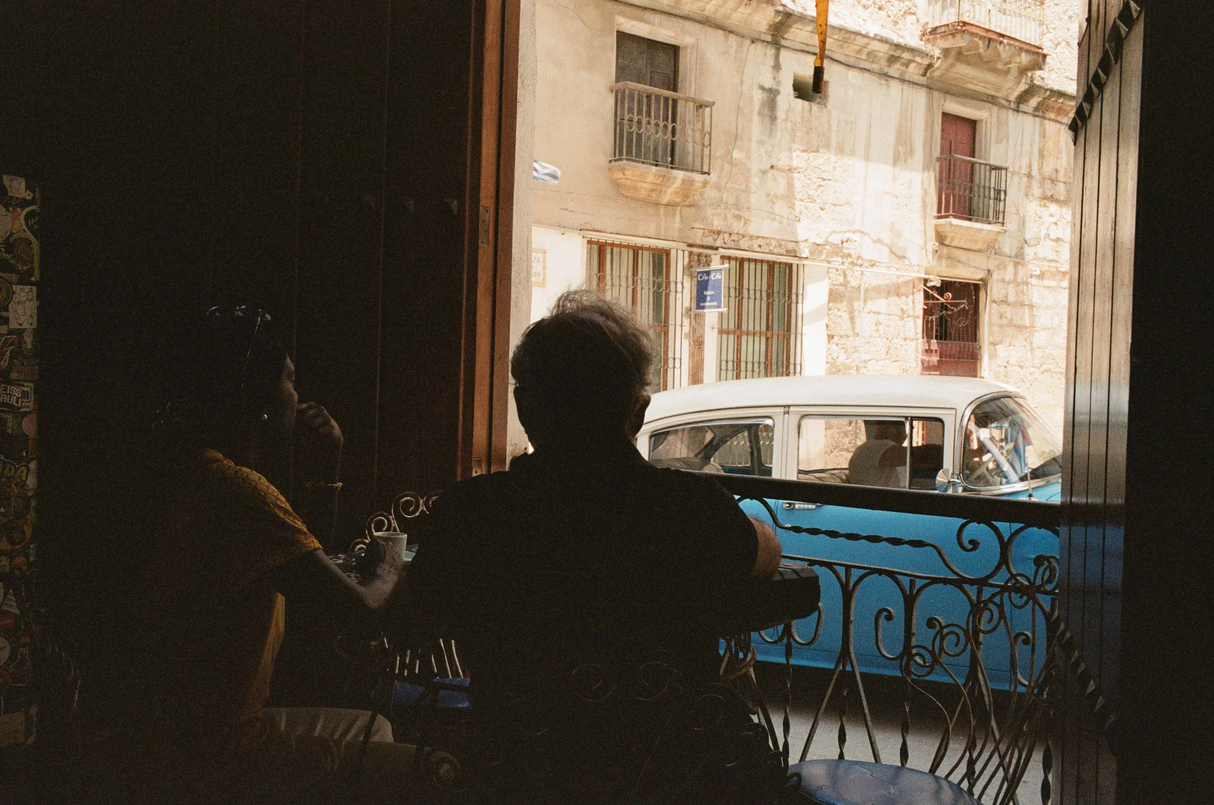 Color film photo of two silhouetted figures at a café table, looking out at a blue vintage car passing on a Havana street.