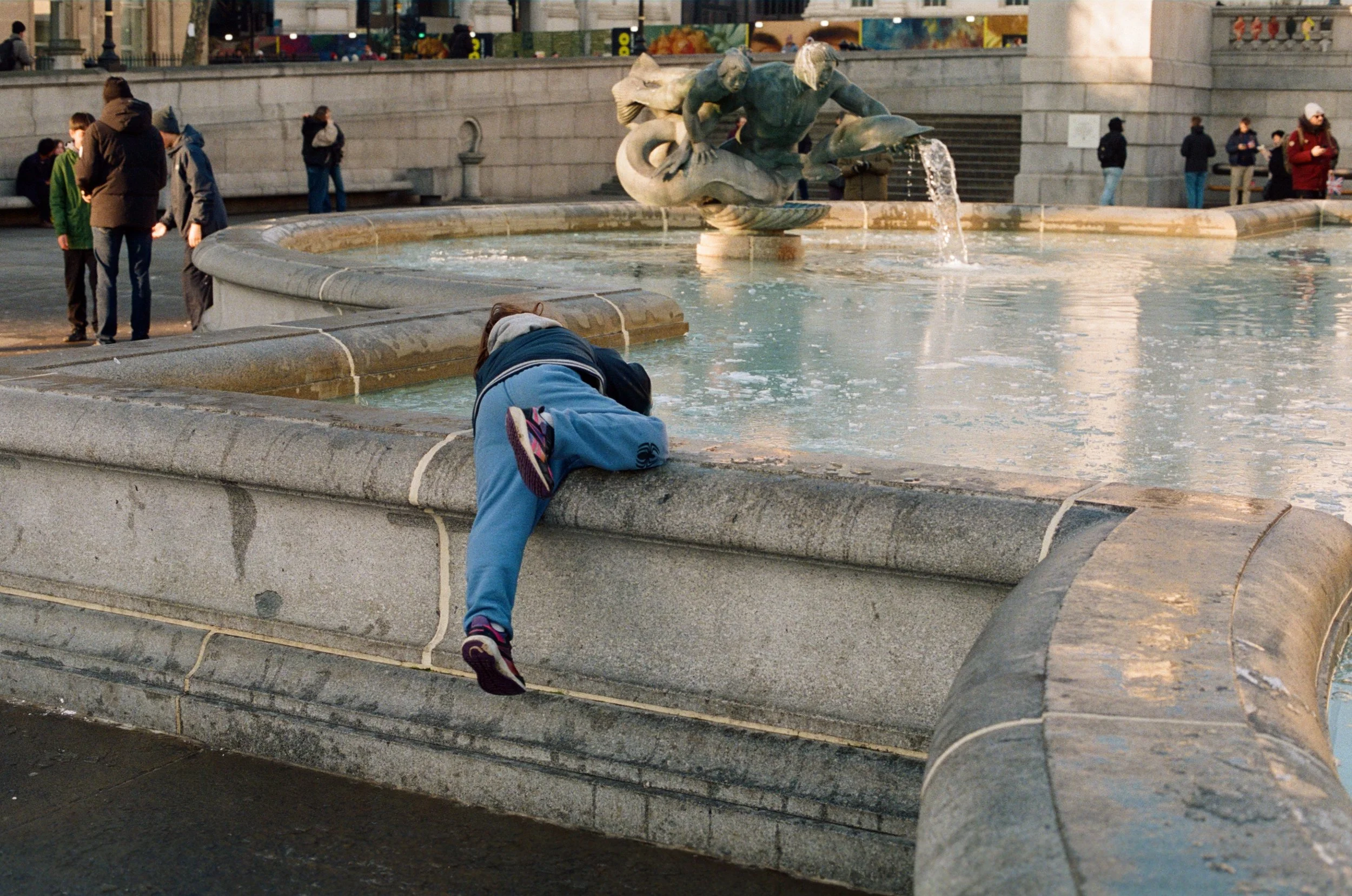 Candid analog film street photograph of a child draped over the stone edge of the Trafalgar Square fountain in London, captured by NYC film photographer Dario Tapia.