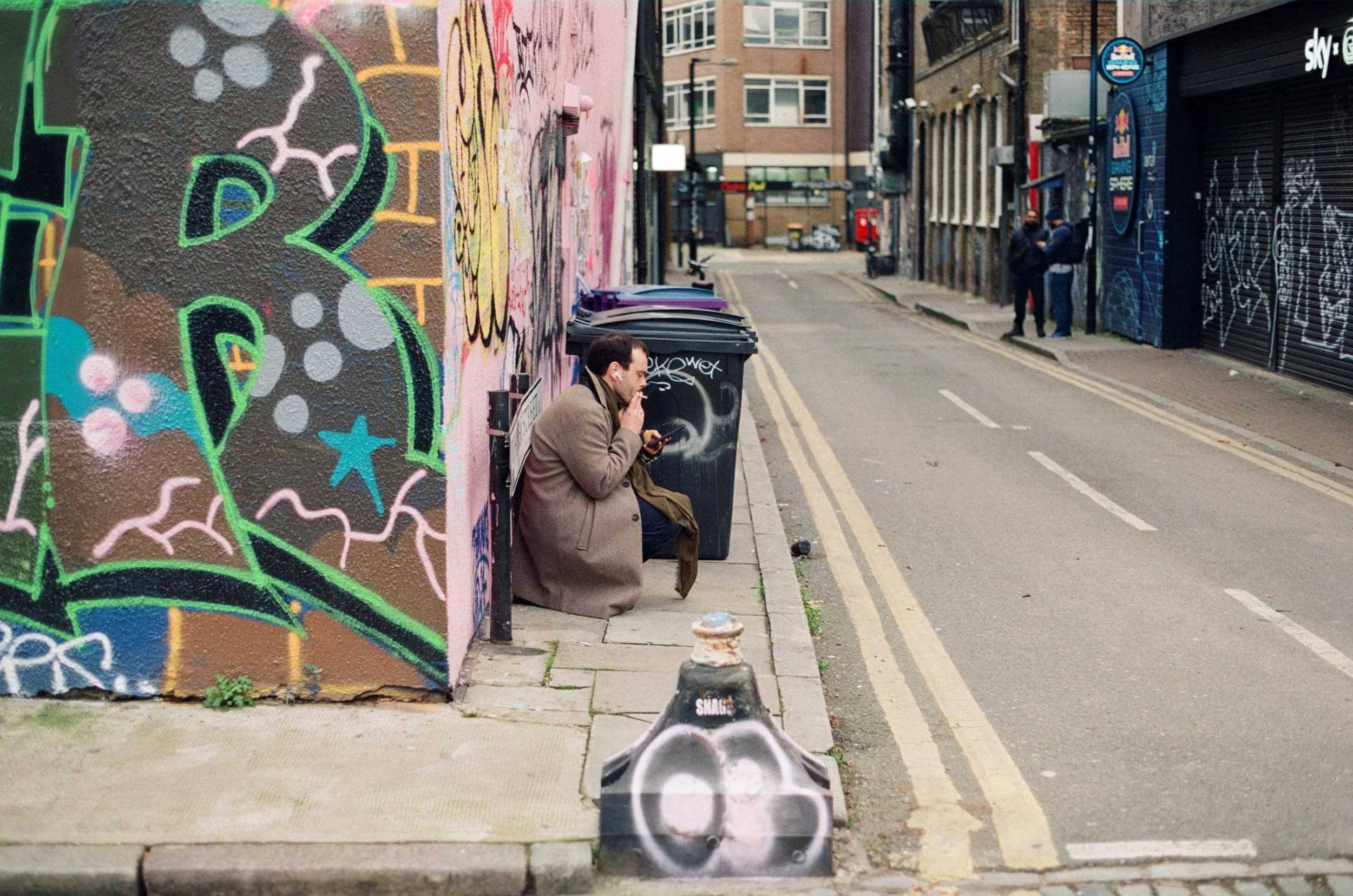 Raw analog film street photograph of a man in a wool coat crouched alone smoking against a graffiti-covered wall in a Shoreditch alley, London, shot by Dario Tapia.