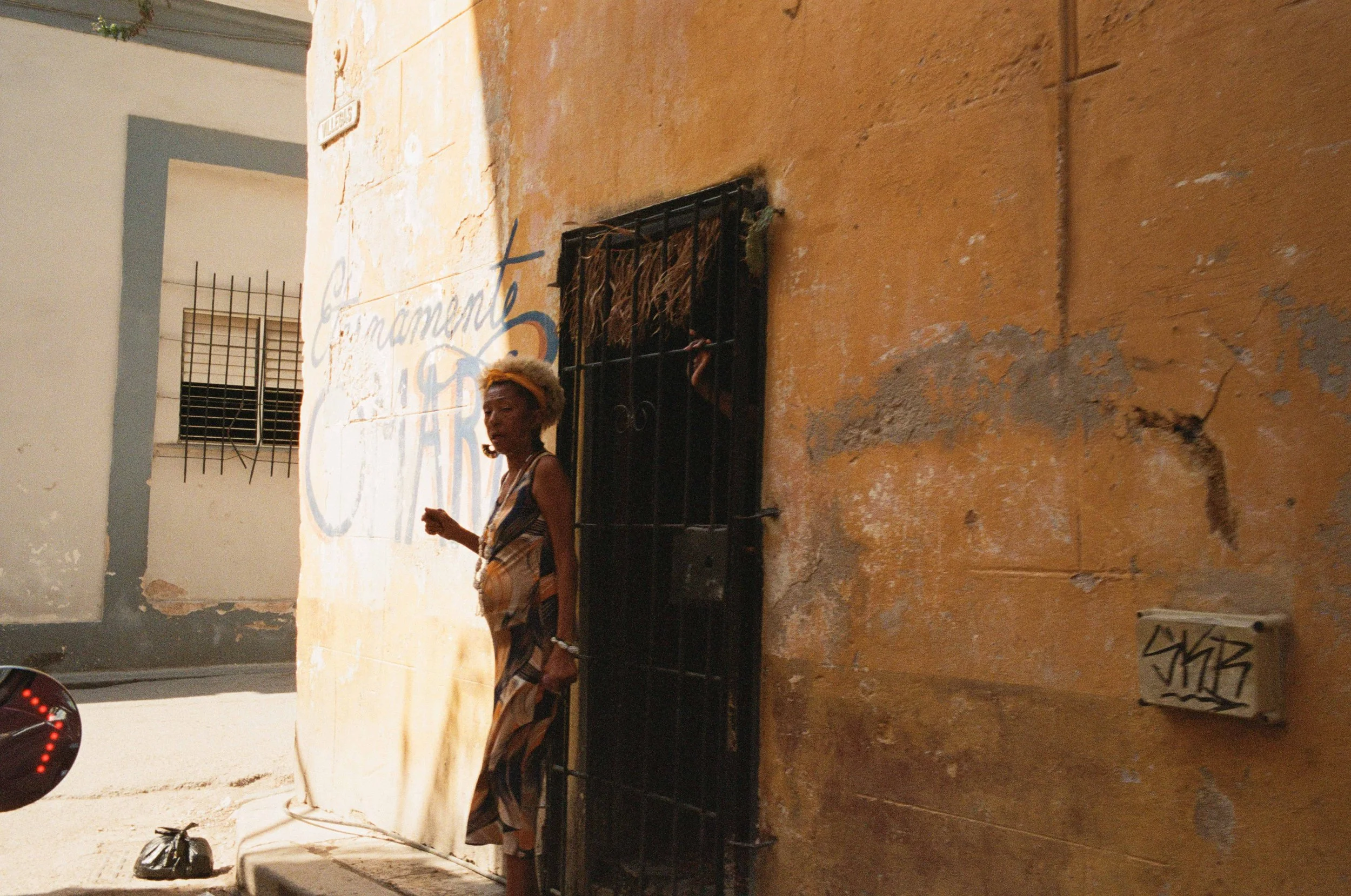 Color film photo of a woman in a patterned dress standing in a sunlit doorway on an ochre-colored street in Havana, Cuba.
