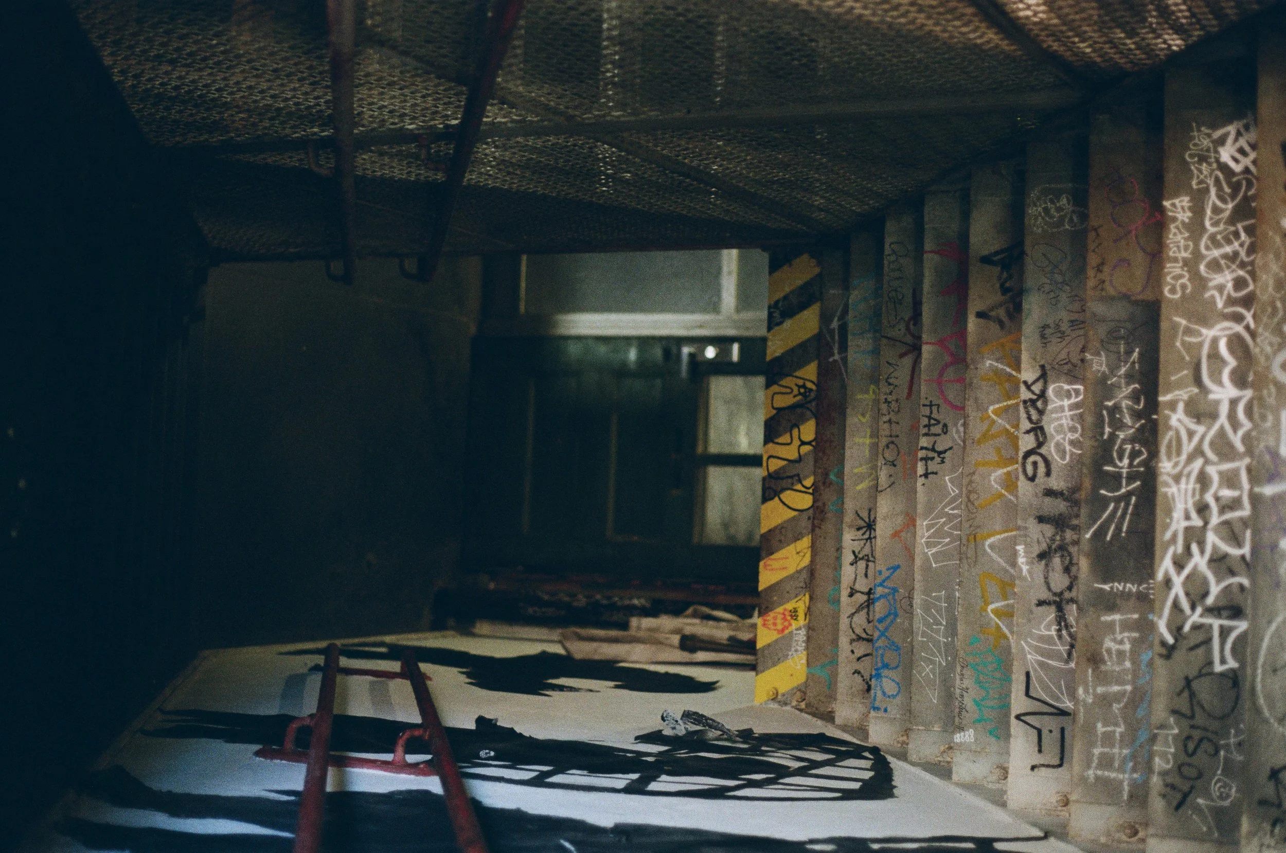 Dark and gritty film street photography of an abandoned urban space — graffiti-covered concrete columns, dramatic shadows, and raw industrial decay captured on film by Dario Tapia, NYC-based street photographer.