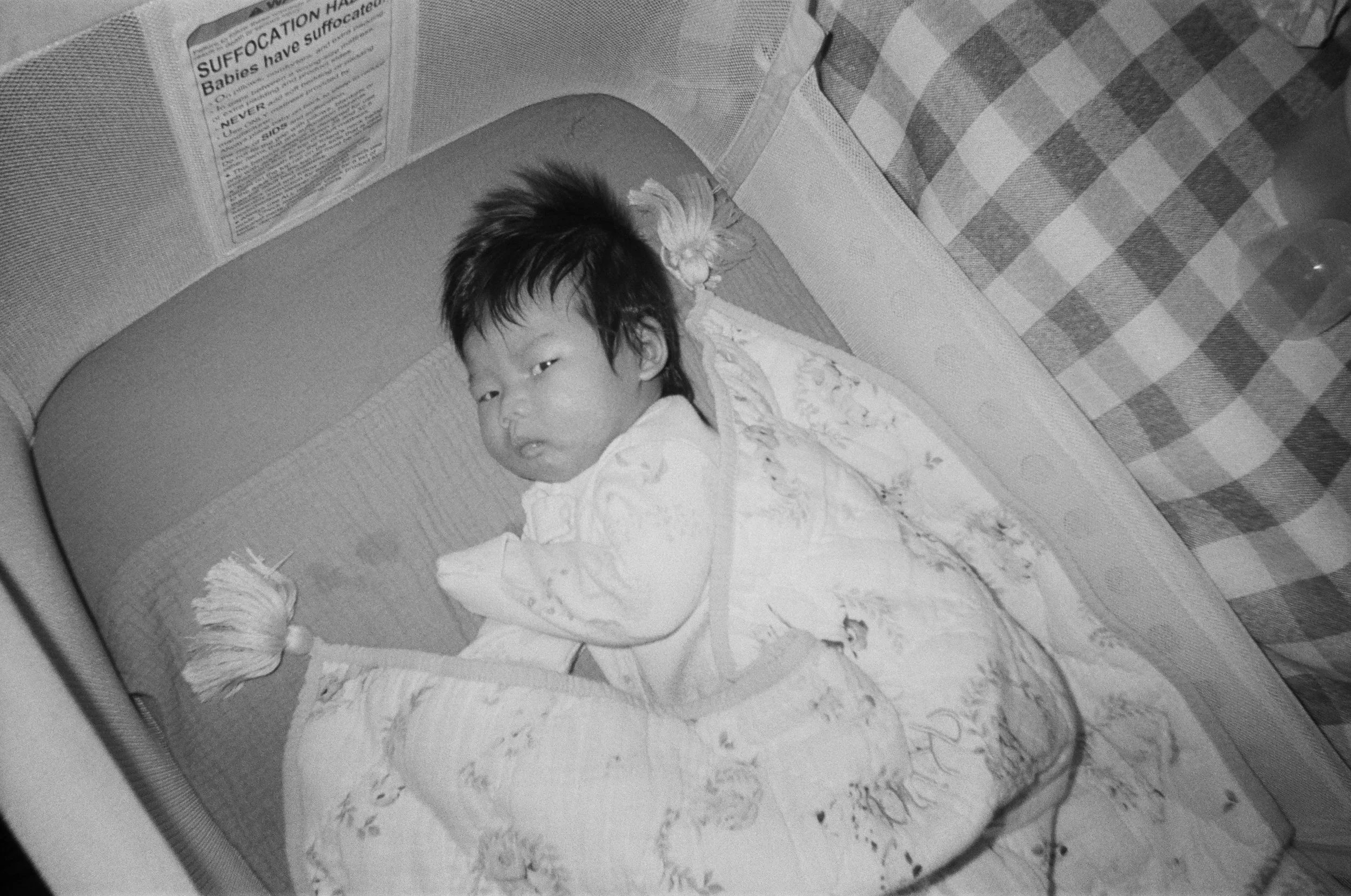 Black and white analog film photograph of a baby with tousled dark hair lying in a crib, looking toward the camera.