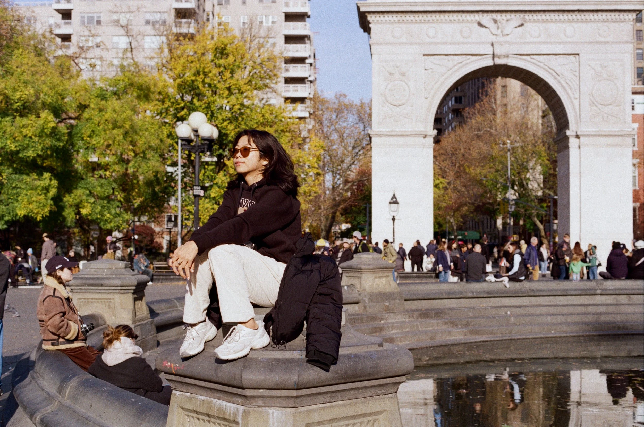 Analog film street photograph of a young woman perched on the Washington Square Park fountain with the iconic arch behind her, captured by NYC film photographer Dario Tapia.