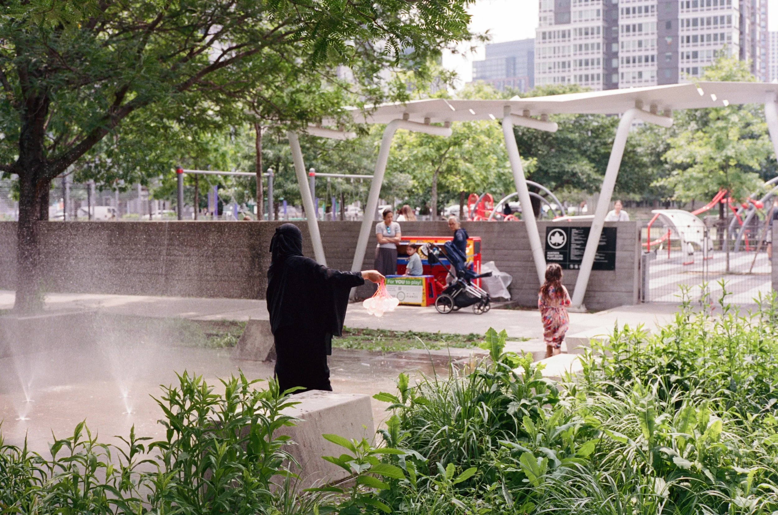 Analog film street photograph of a woman in a black niqab reaching toward a fountain spray in a New York City park, captured by film photographer Dario Tapia.