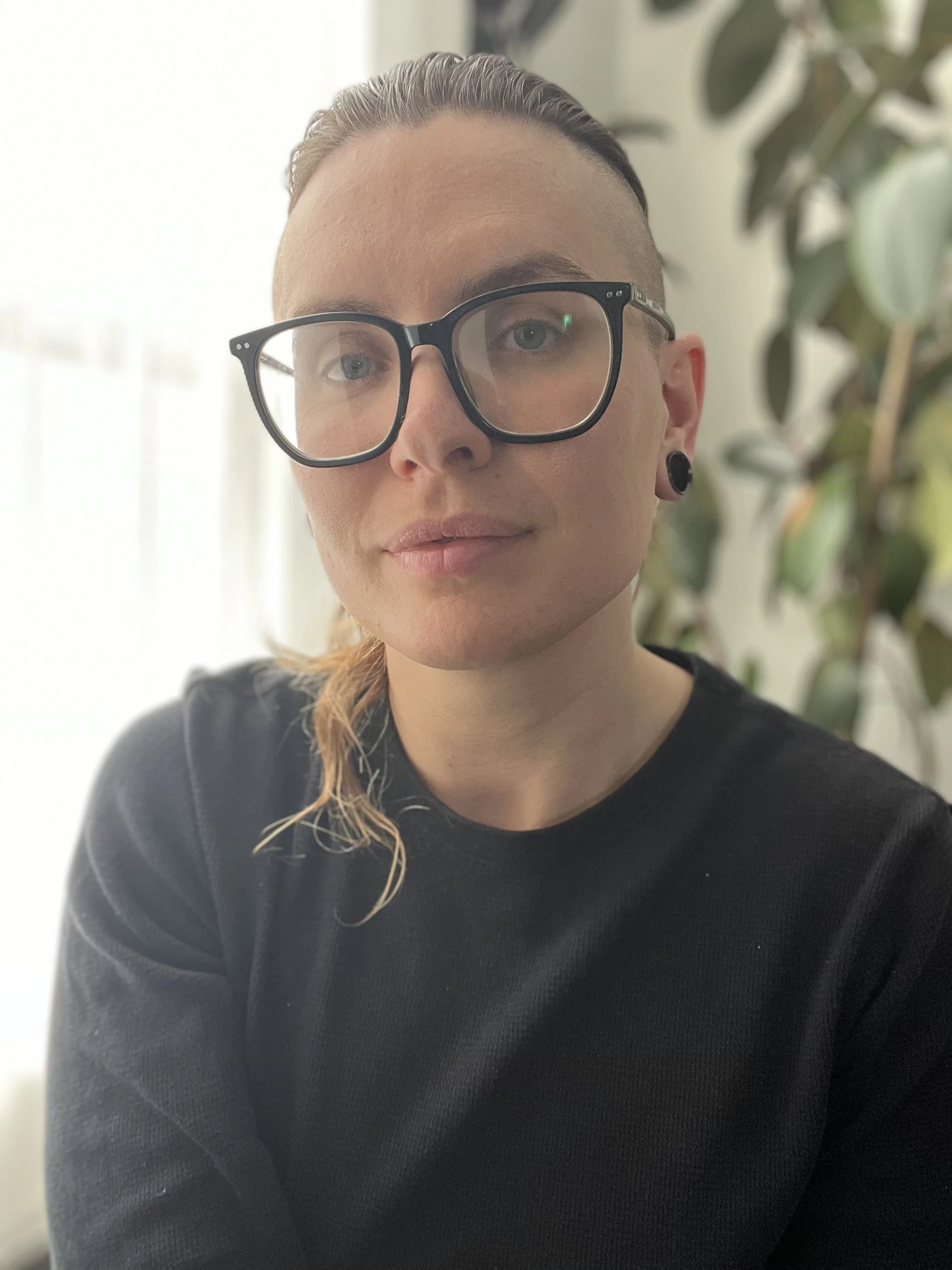 Close-up portrait of a person with short hair, wearing large black glasses, black earrings, and a black shirt, posing indoors in front of a leafy plant and a bright window.