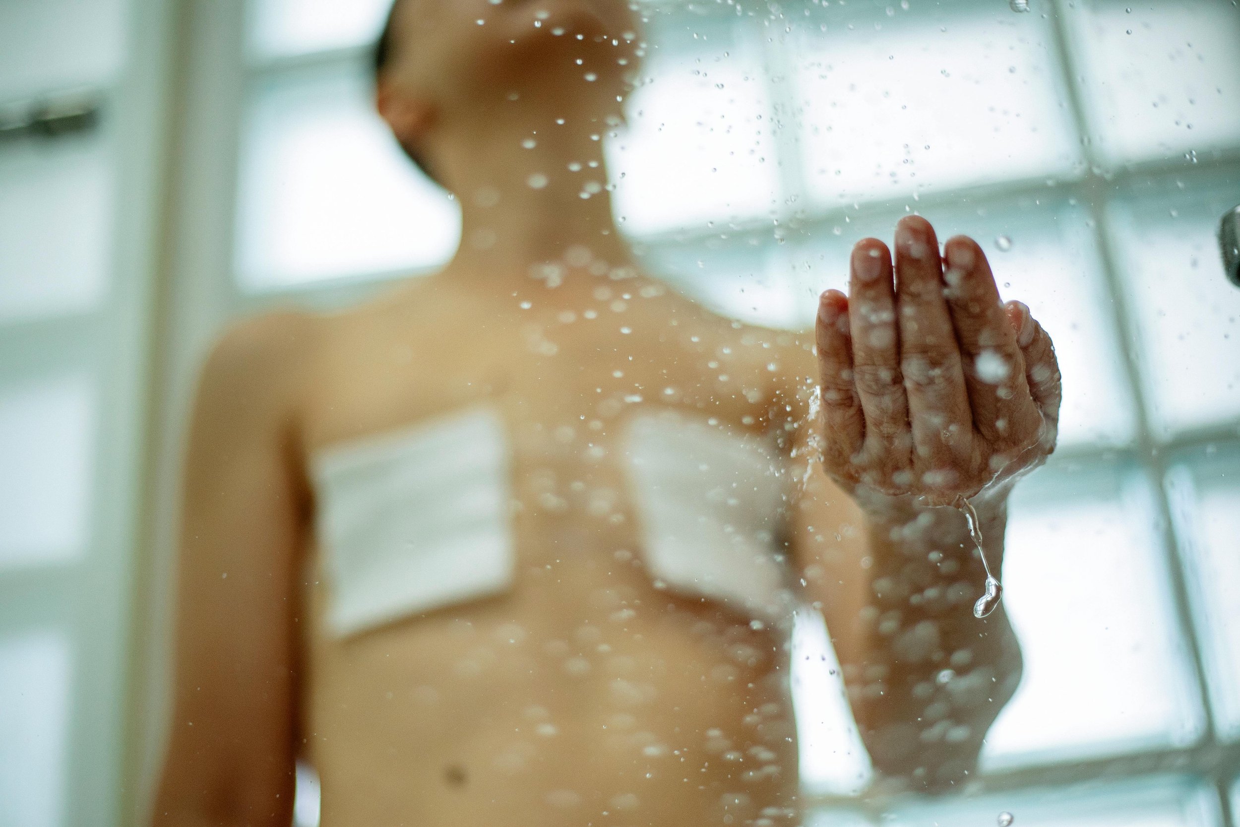 A person standing behind a foggy glass shower door, with their hand pressed against the glass. The scene appears to be taken in a bathroom with a window in the background. They appear to have recently undergone top surgery.