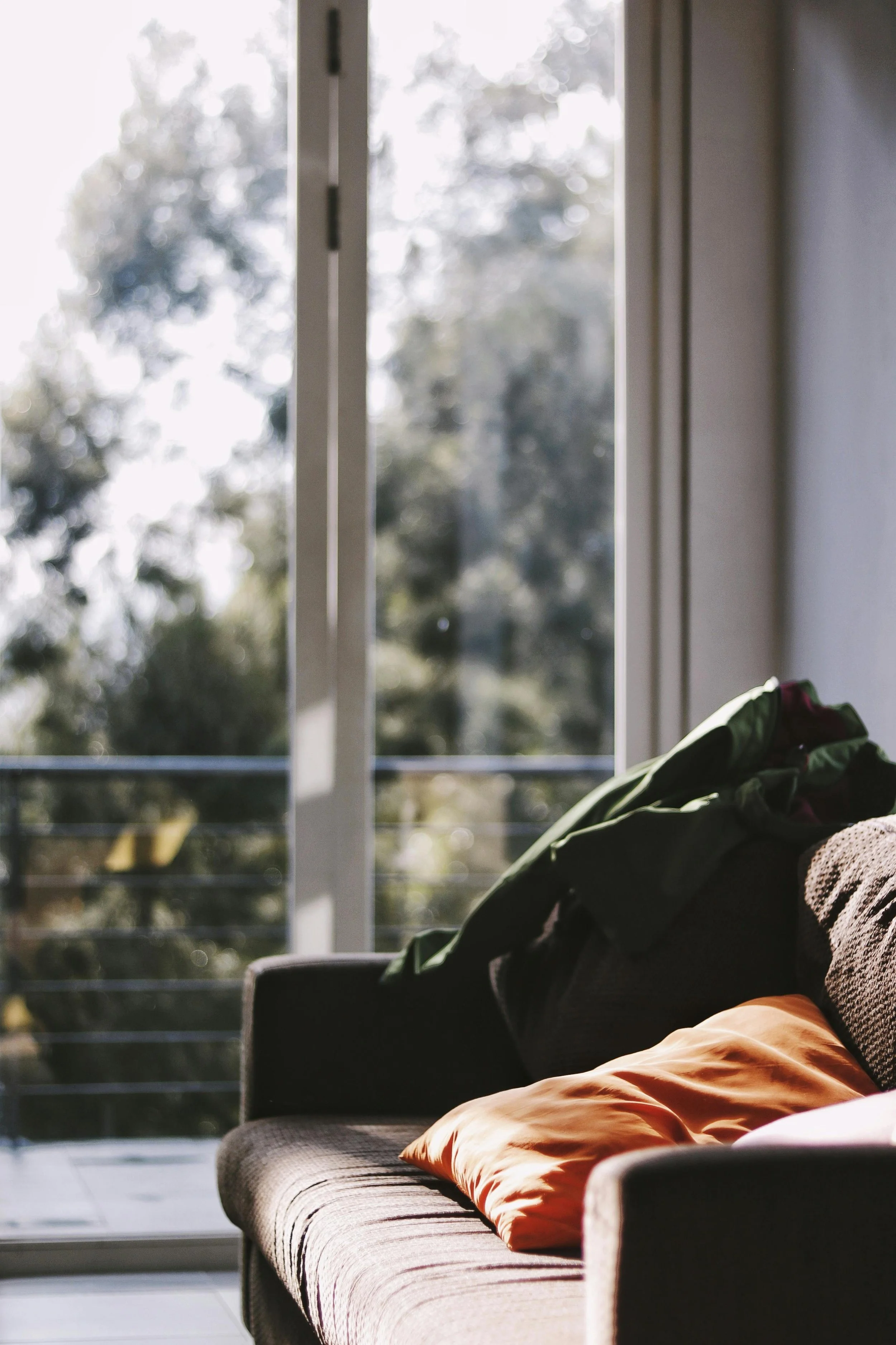 A sofa with a pillow and a folded blanket inside a room, near a large sliding glass door that opens to a balcony with a metal railing, with trees visible outside in the background.