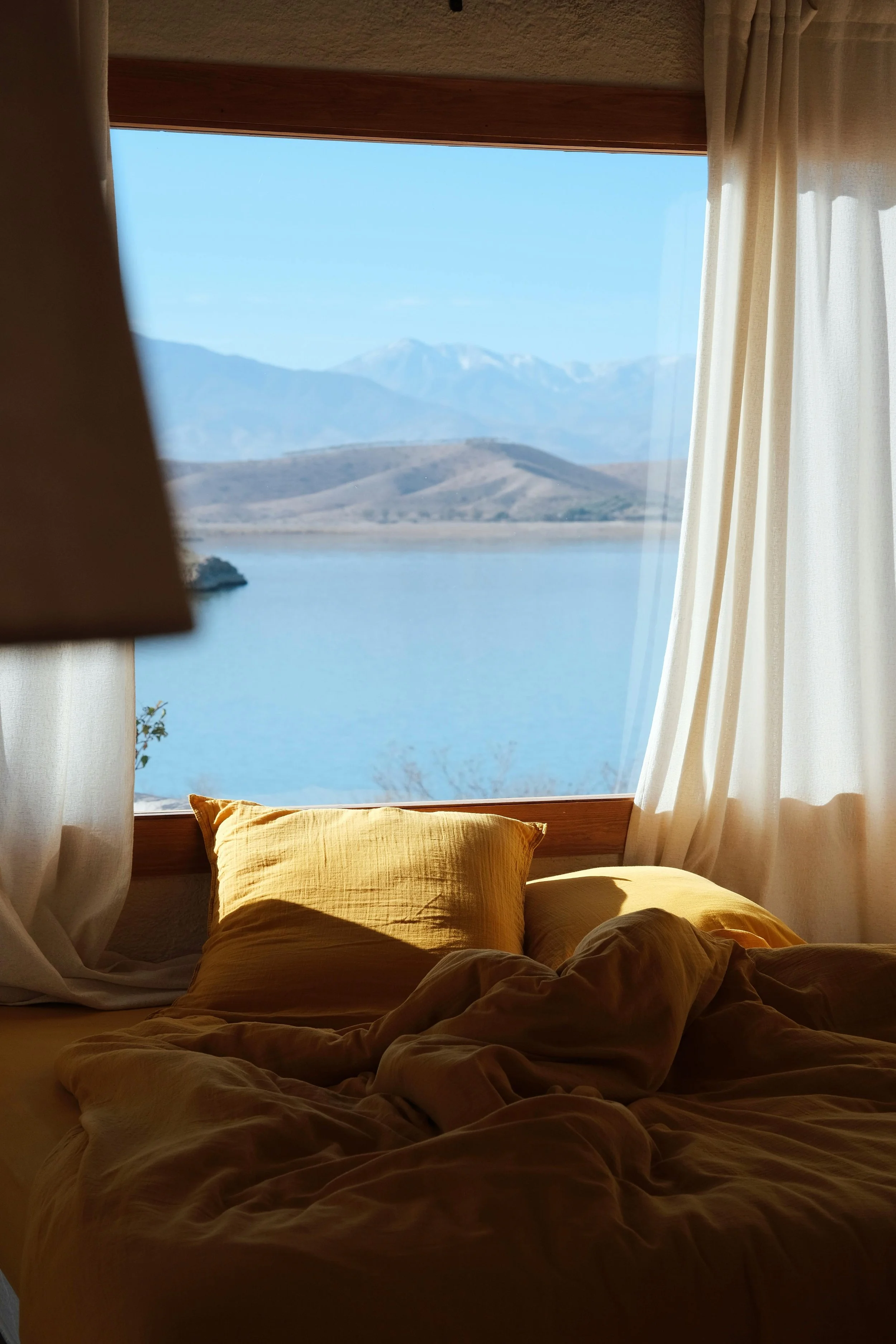 A bedroom with a messy bed and mustard-colored pillows, looking out a large window at a scenic landscape with a lake, rolling hills, and snow-capped mountains in the distance, with white curtains framing the view.
