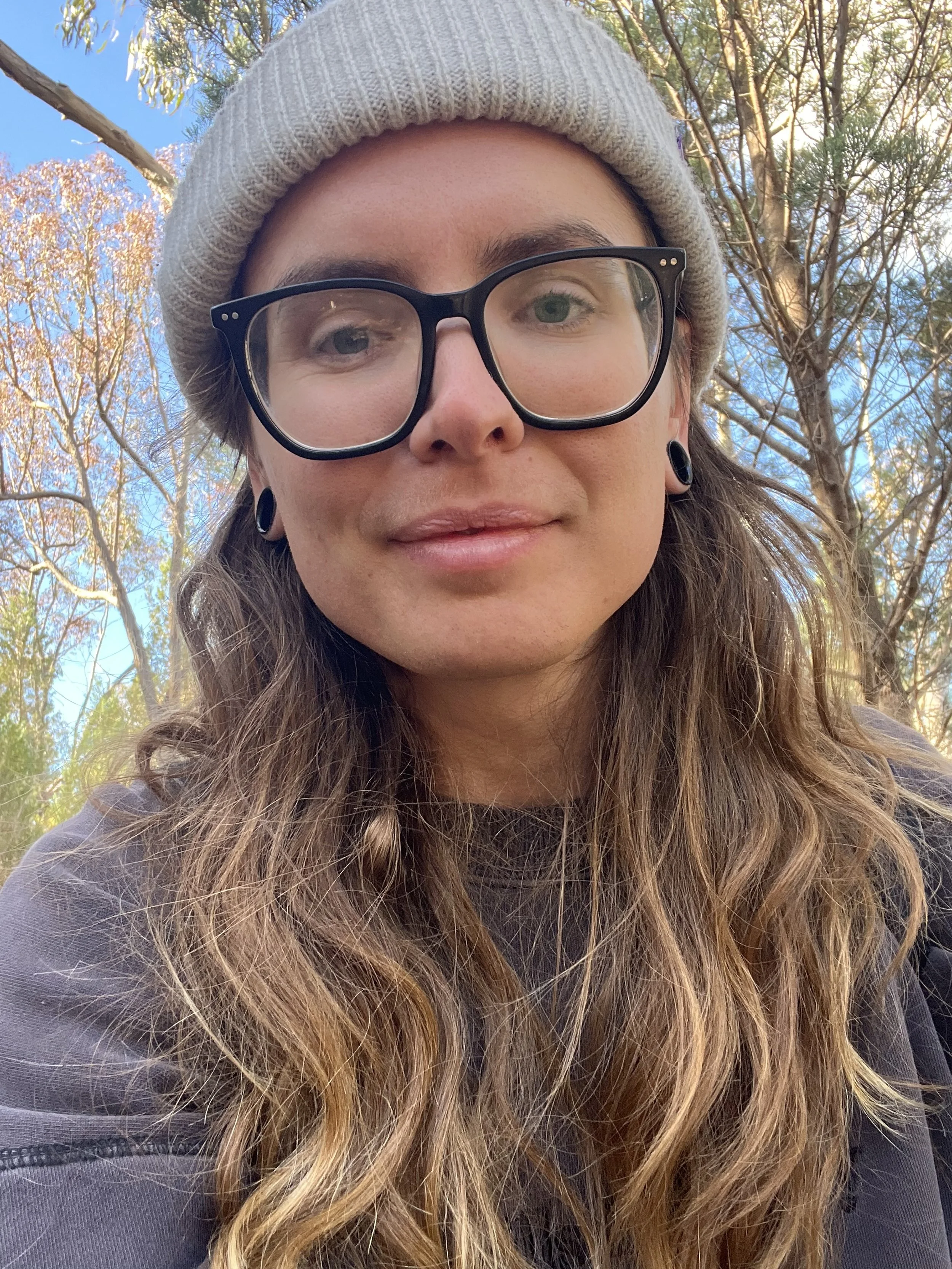 Close-up of a person with long wavy hair, wearing black glasses, a beige beanie, and black earrings, outdoors with trees and blue sky in the background.