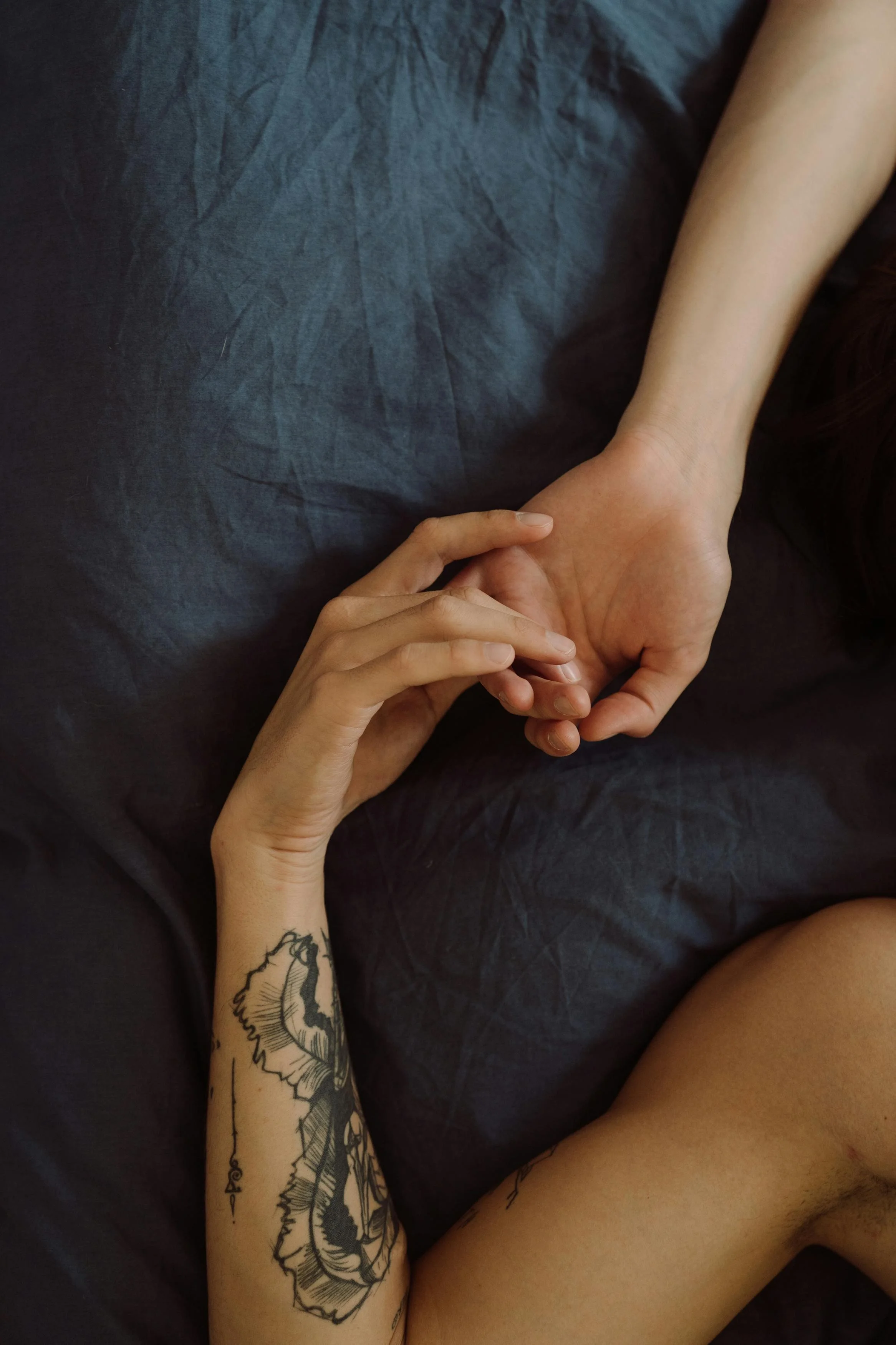 Close-up of two people holding hands, one with a tattooed arm, lying on a dark surface.