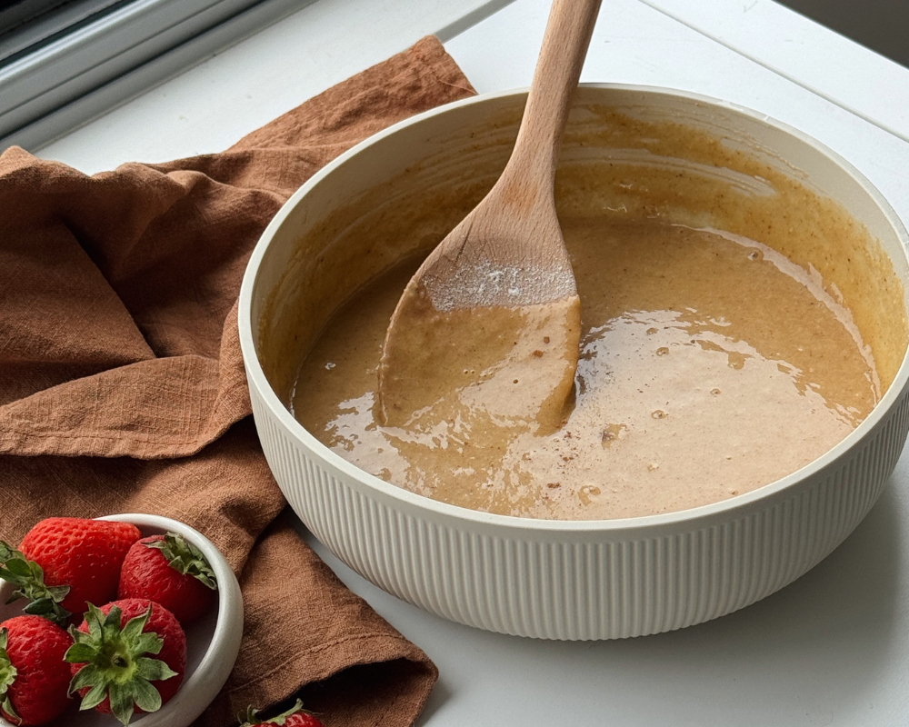 A bowl of cake batter with a wooden spoon and small bowl of strawberries next to it