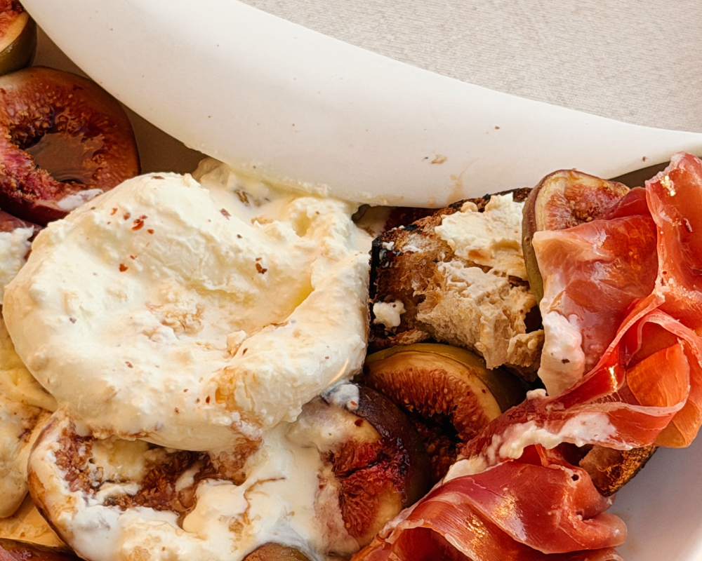  Close up of burst burrata with creamy filling spilling over fresh figs, prosciutto and toasted sourdough 