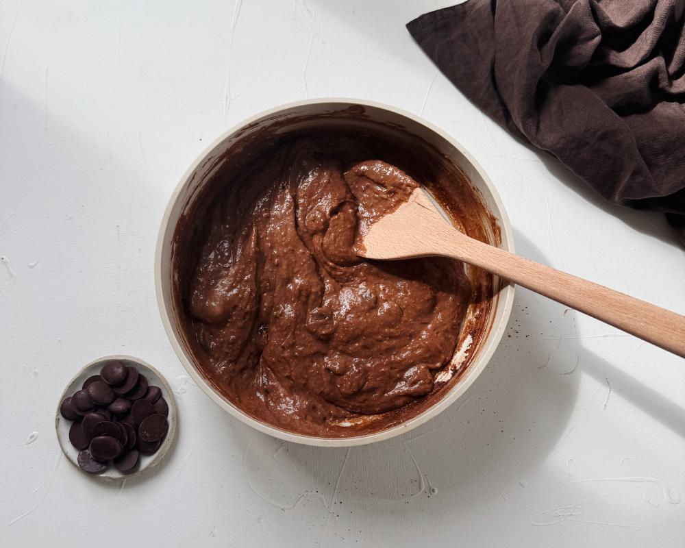  Rich chocolate cake batter in a ceramic bowl with a wooden spoon, dark chocolate buttons on the side 