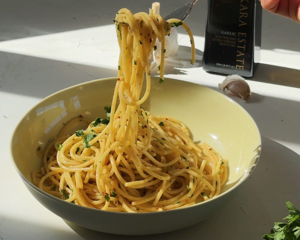  A twirl of spaghetti aglio e olio lifted with a fork over a bowl, with Pukara Estate olive oil in the background 