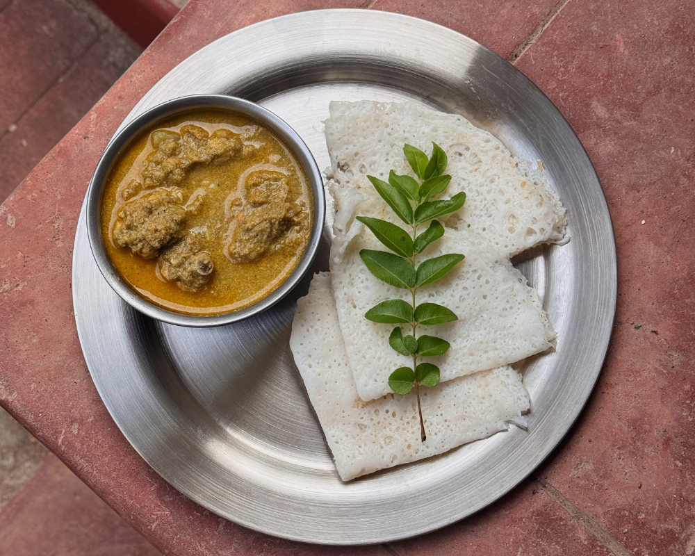  A steel plate with a bowl of Mangalorean green chicken curry served alongside folded neer dosa, garnished fresh curry leaves 