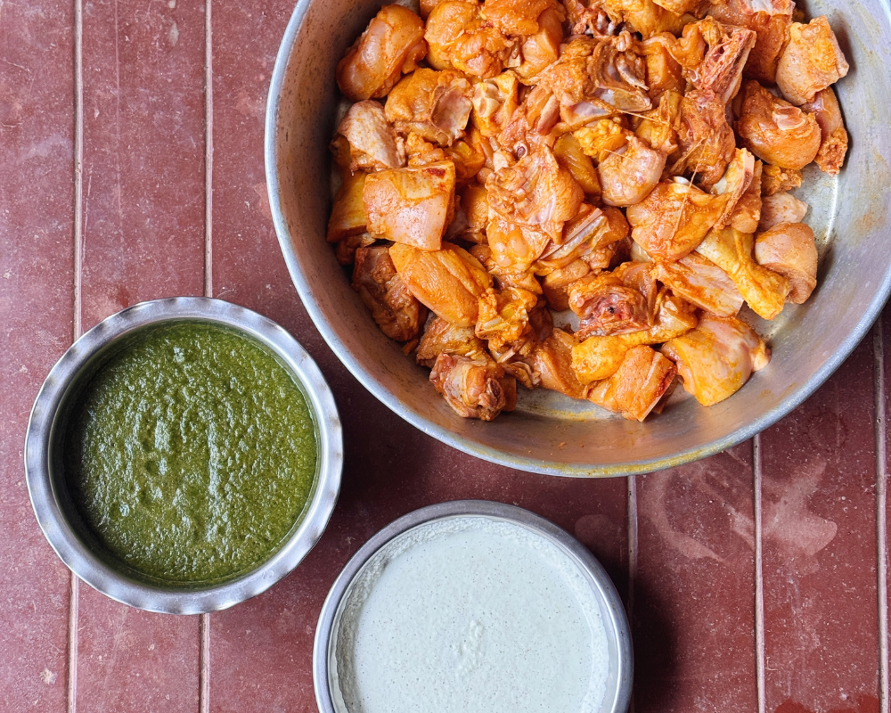 Three steel bowls on a red tiled surface — marinated bone-in chicken pieces, a vibrant green herb paste, and coconut cream, ready to be combined for Mangalorean green chicken curry.