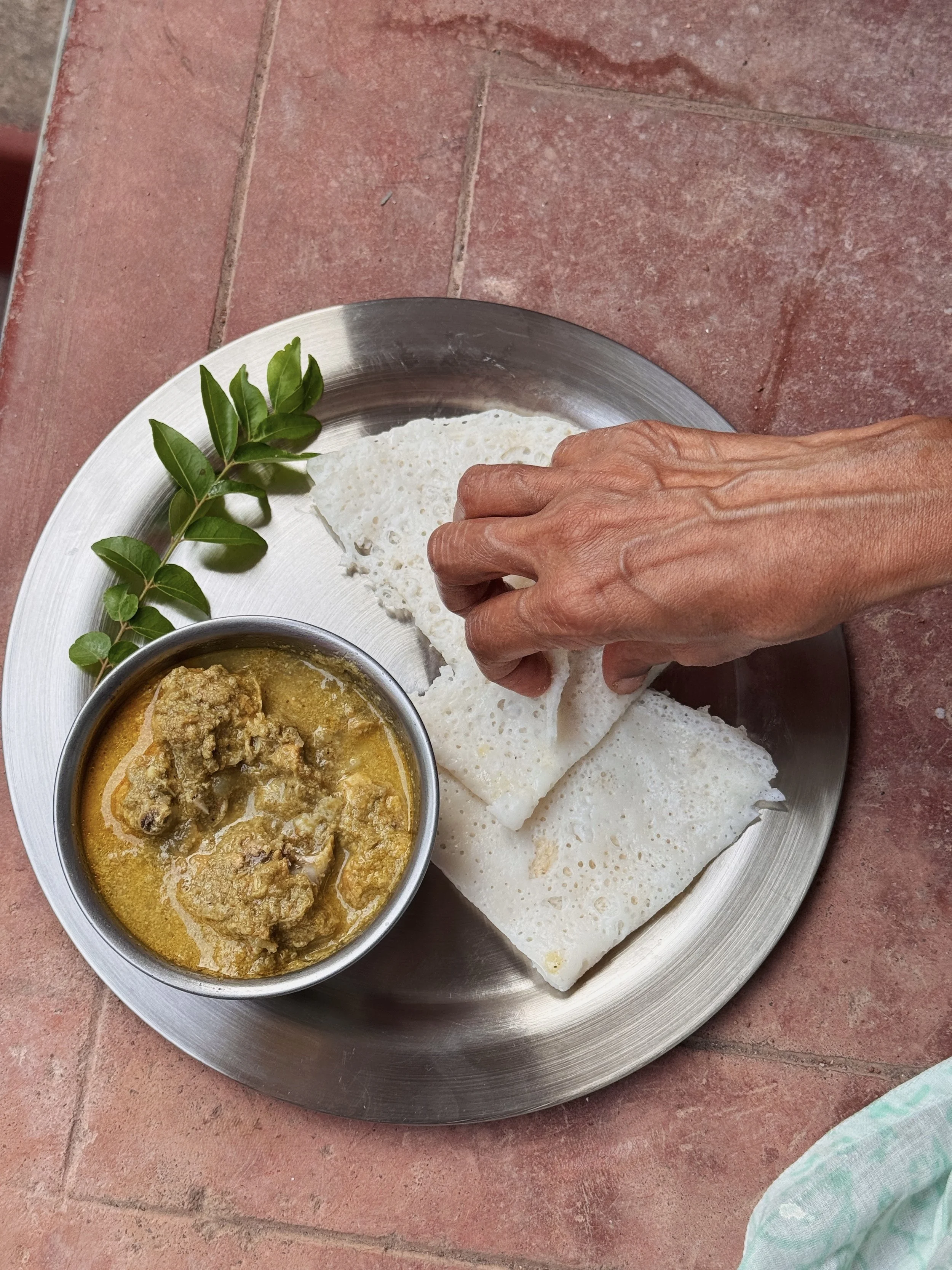  A hand reaching into a steel plate with a bowl of Mangalorean green chicken curry served alongside folded neer dosa, garnished fresh curry leaves 