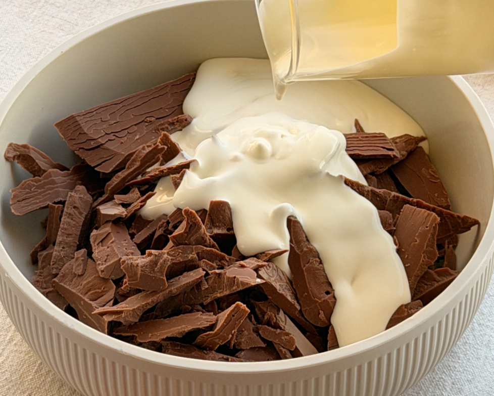  Cream being poured over broken Gianduja chocolate pieces in a white ceramic bowl 