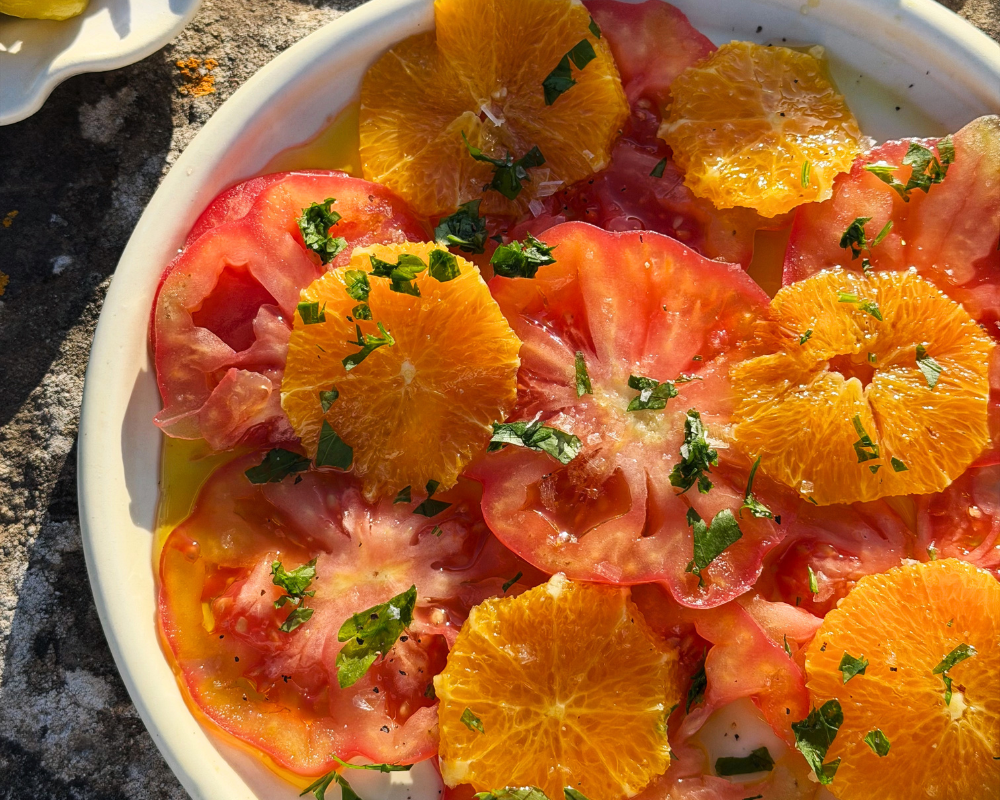  A close-up overhead shot of a tomato and orange salad in a white ceramic dish, topped with chopped fresh parsley and a golden olive oil dressing, shot in natural sunlight. 