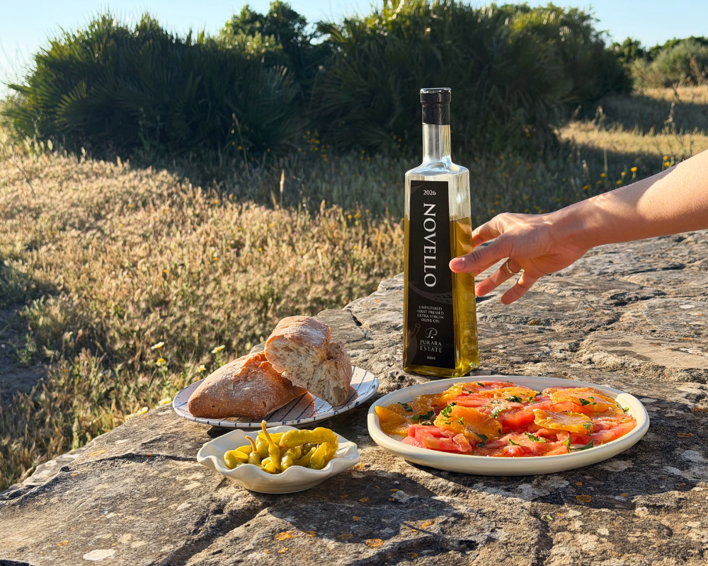  A rustic outdoor spread on a stone surface featuring a plate of tomato and orange salad, torn crusty bread, a small dish of pickled peppers and a bottle of Pukara Estate Novello extra virgin olive oil, with a hand reaching in and wild grass in the b