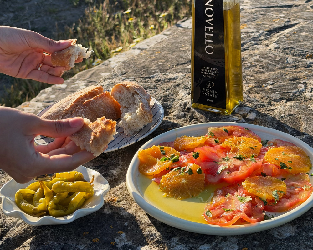  Two hands tearing a chunk of crusty bread beside a plate of tomato and orange salad and a bottle of Pukara Estate Novello olive oil on a stone ledge, with a small dish of pickled peppers and golden afternoon light in the background. 