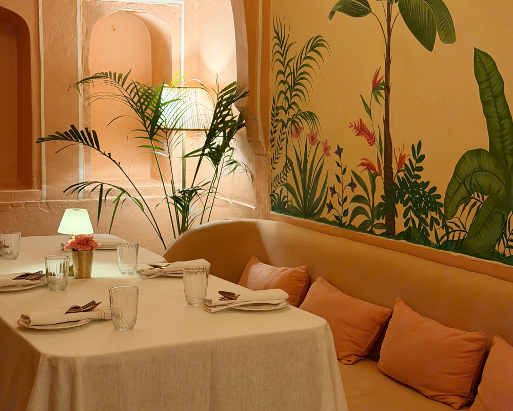 A cosy dining nook with blush banquette seating and a tropical mural at The Johri Restaurant, Jaipur. 