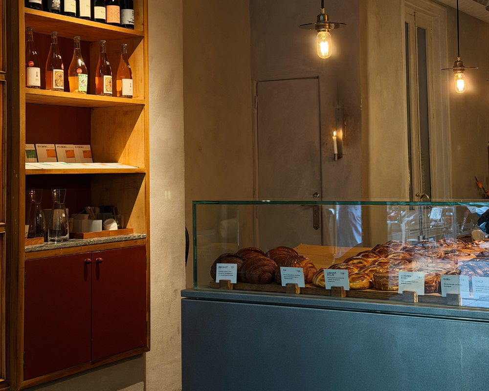  Acid Cafe’s counter displaying croissants and pastries under glass, with natural wine bottles on wooden shelves behind. 