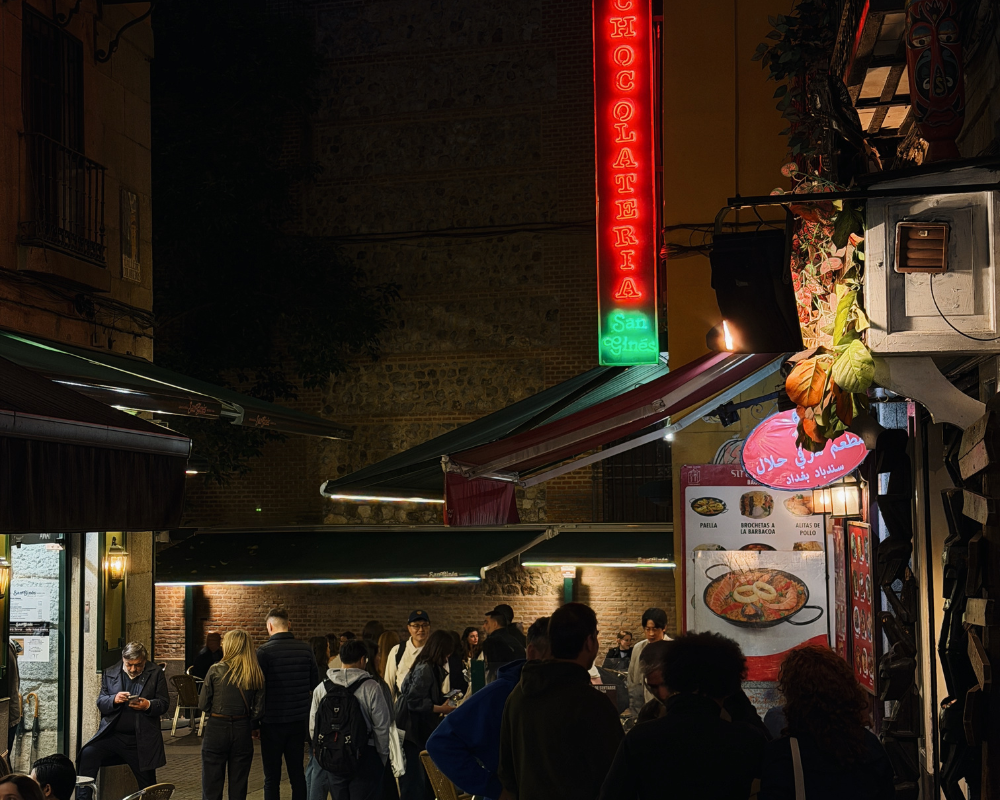  The neon sign of Chocolatería San Ginés glowing red at night in a busy Madrid alleyway. 