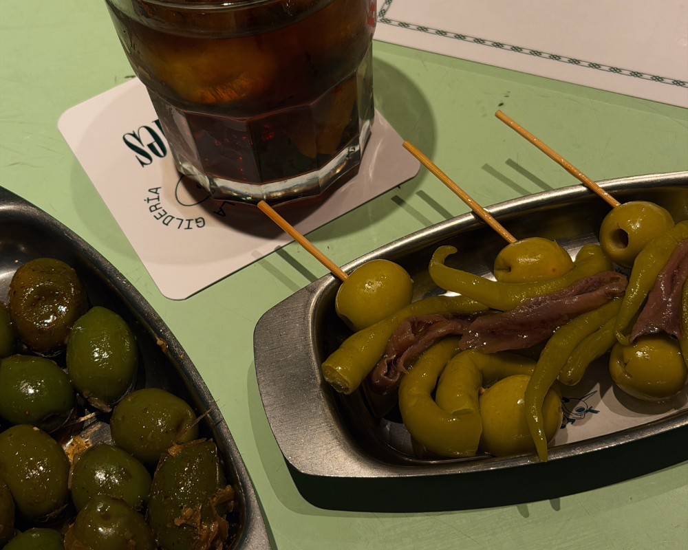  Small dishes of gildas with green olives and anchovies on skewers, alongside a glass of vermouth at La Gilderia Madrid 