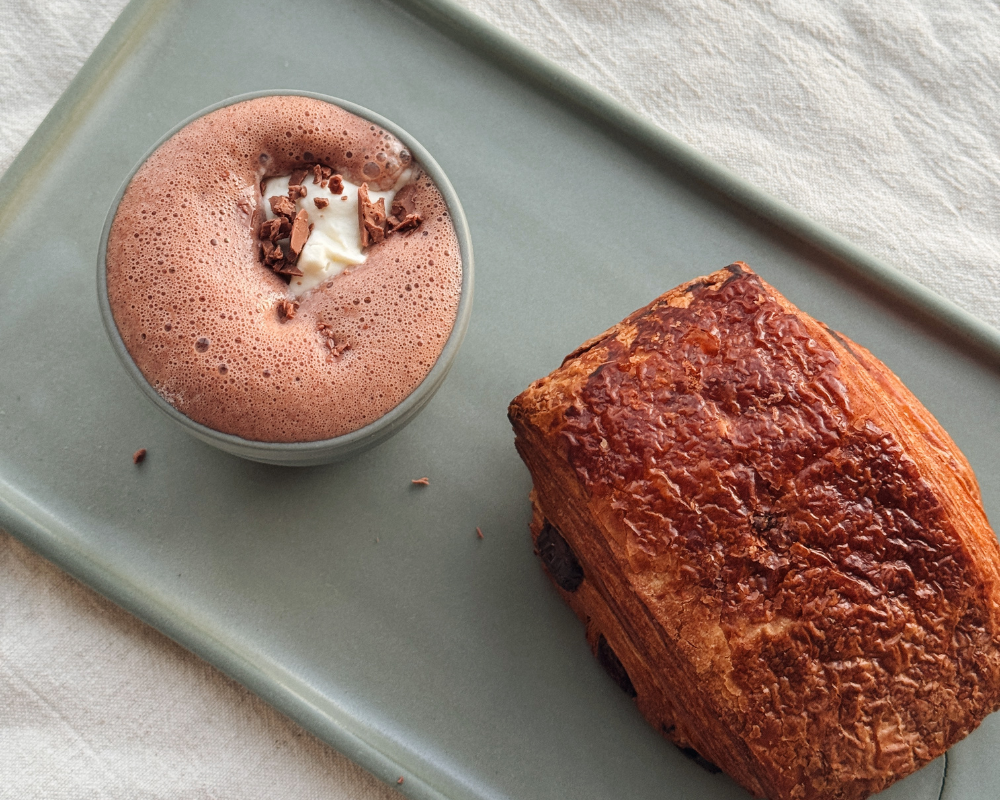  A cup of Chai Hot Chocolate next to a pain au chocolat on a grey plate 
