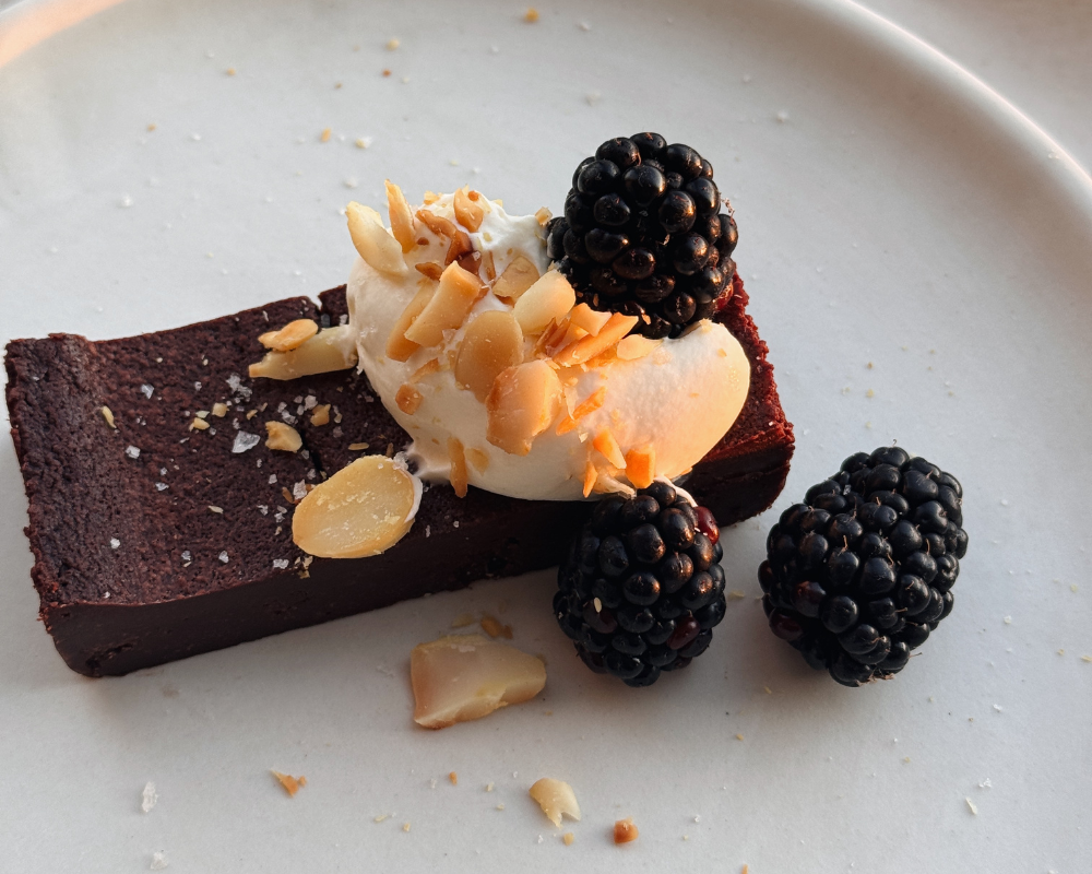  Slice of baked chocolate ganache with blackberries, cream and toasted nuts on a white plate 