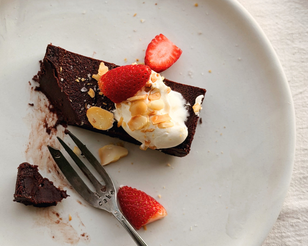  Slice of baked chocolate ganache topped with strawberries, toasted macadamias and cream on a white plate with a fork next to it 