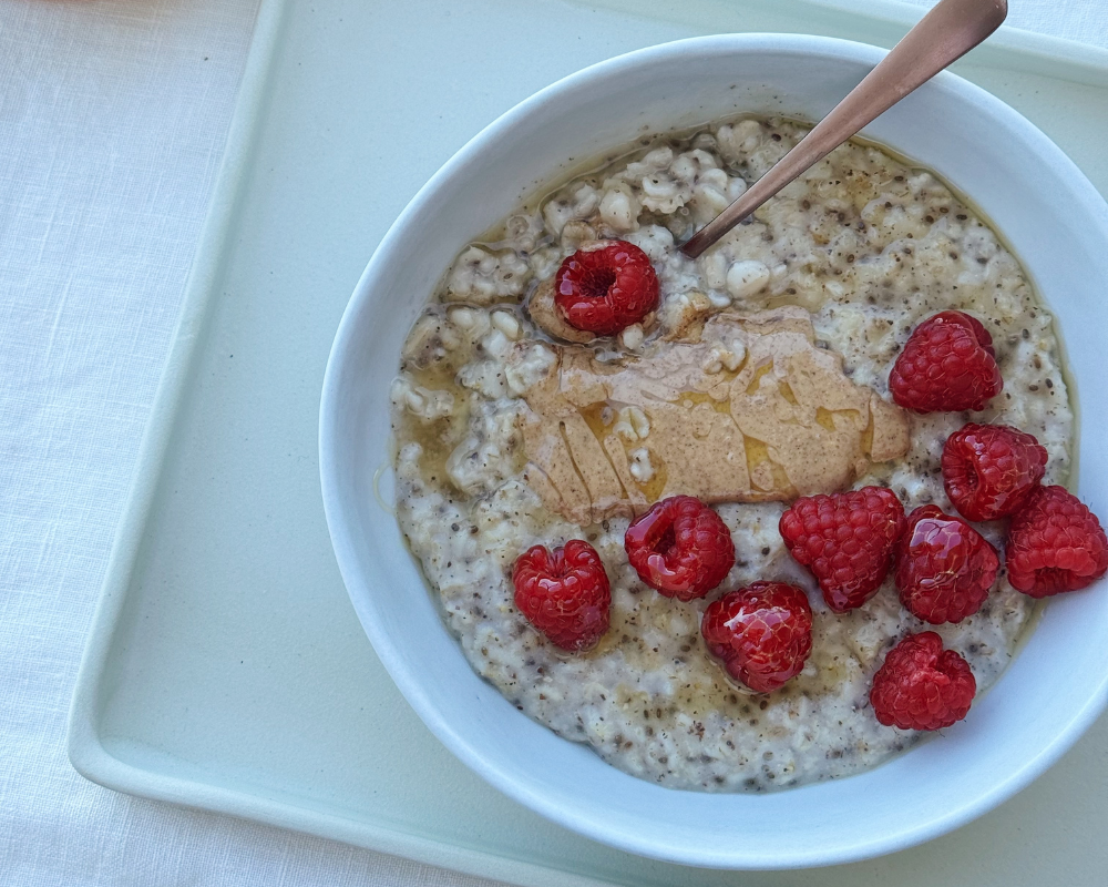  Overhead view of a bowl of creamy oatmeal topped with a swirl of nut butter, honey drizzle, chia seeds, and fresh raspberries, with a spoon resting in the bowl on a light blue tray. 