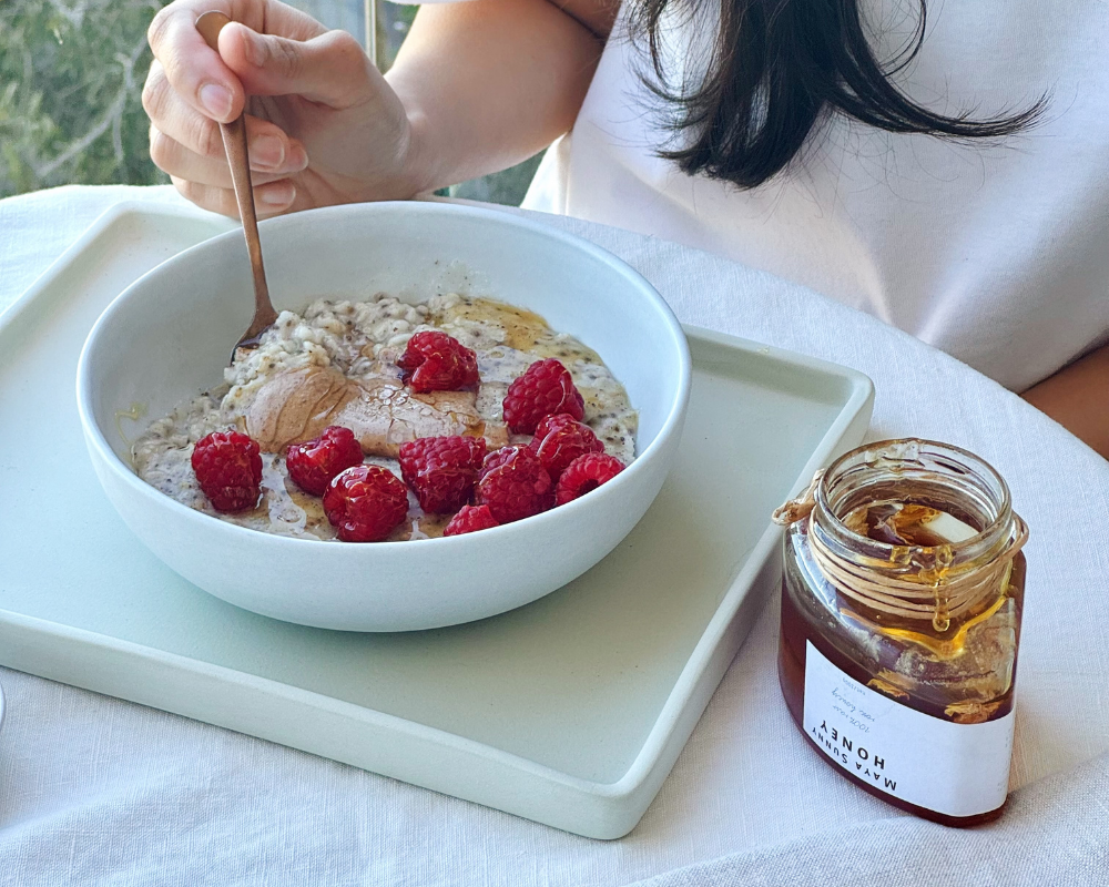 A person's hand stirring a bowl of oatmeal topped with raspberries and nut butter with a spoon, seated at a table next to an open jar of honey