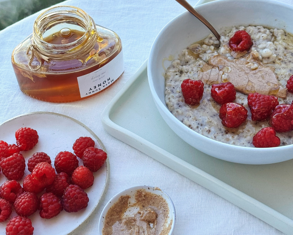  A bowl of oatmeal topped with raspberries and nut butter sits on a tray beside an open honey jar, a small plate of fresh raspberries, and a dish of nut butter. 